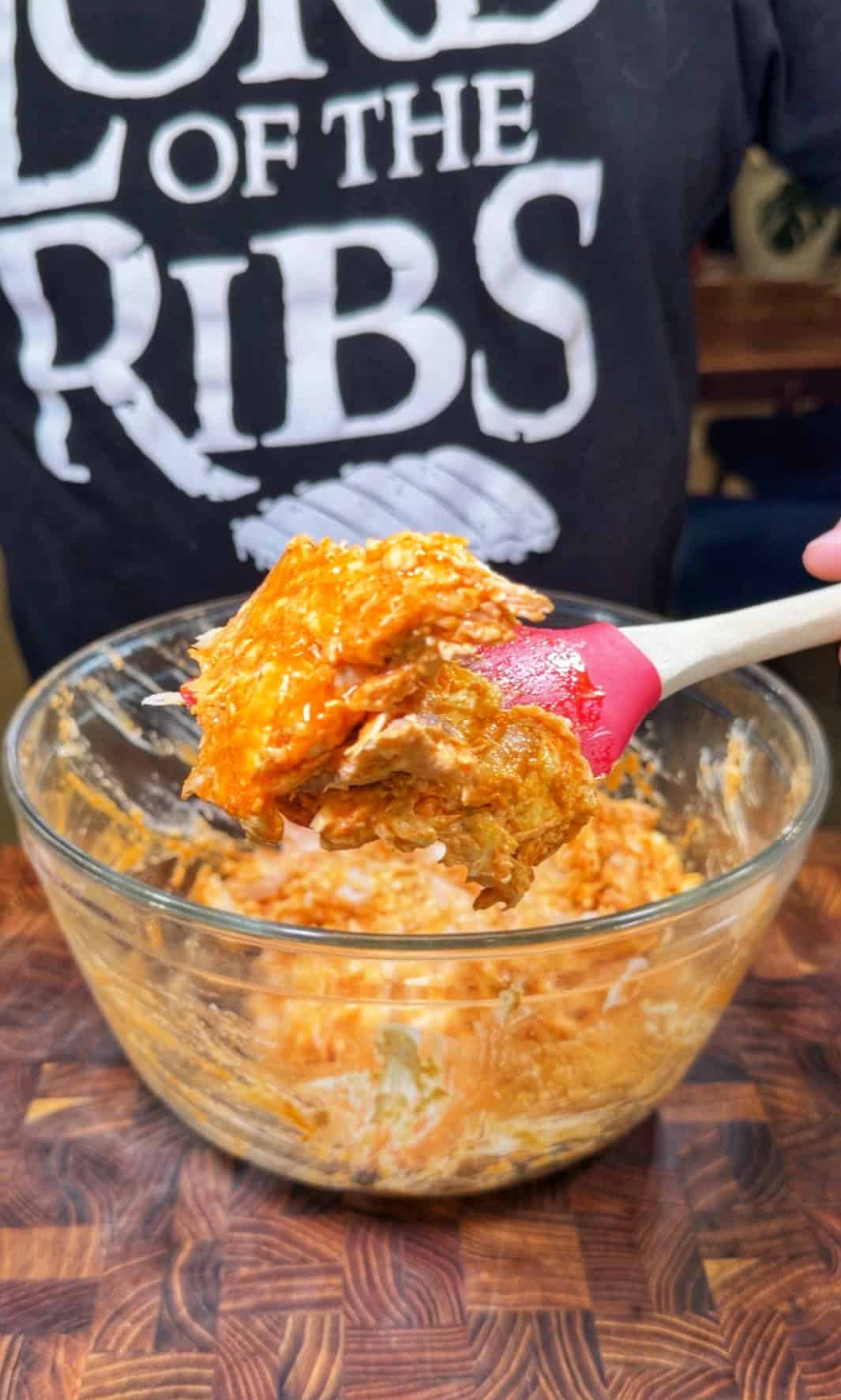 A person wearing a Lord of the Ribs shirt holds a spatula with saucy shredded meat above a glass bowl filled with the same mixture, on a wooden surface.