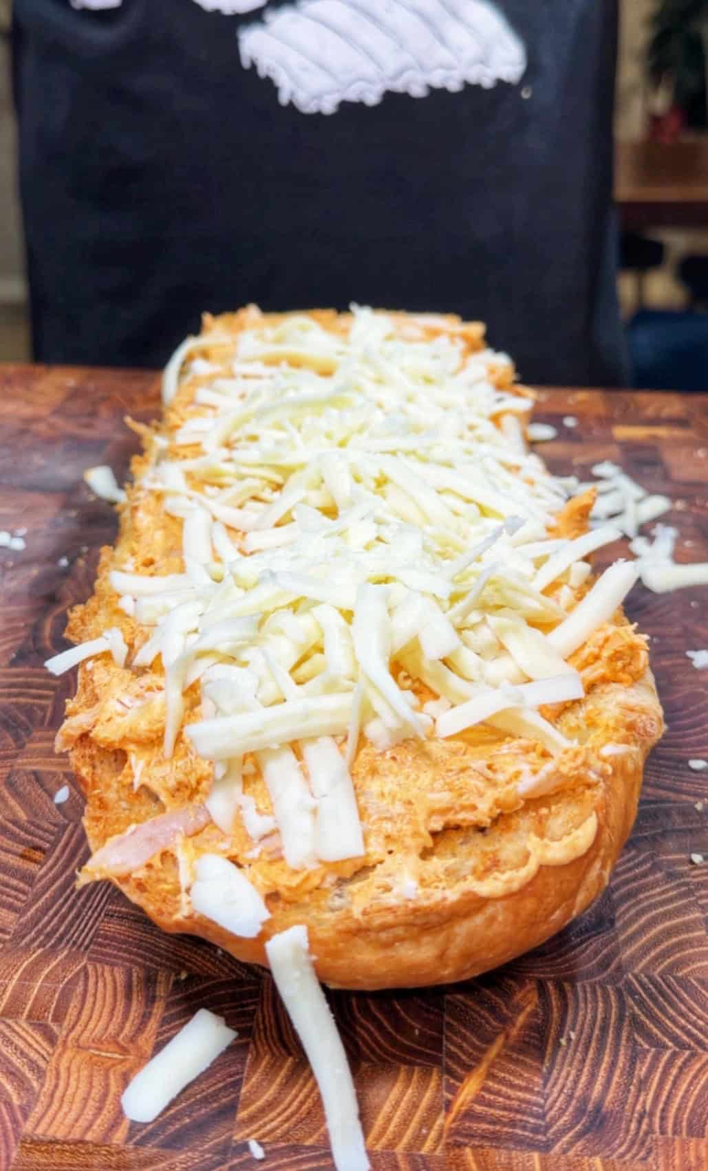 A sliced loaf of bread topped with a layer of shredded cheese sits on a wooden cutting board, ready to be baked. A person wearing a dark apron is visible in the background.