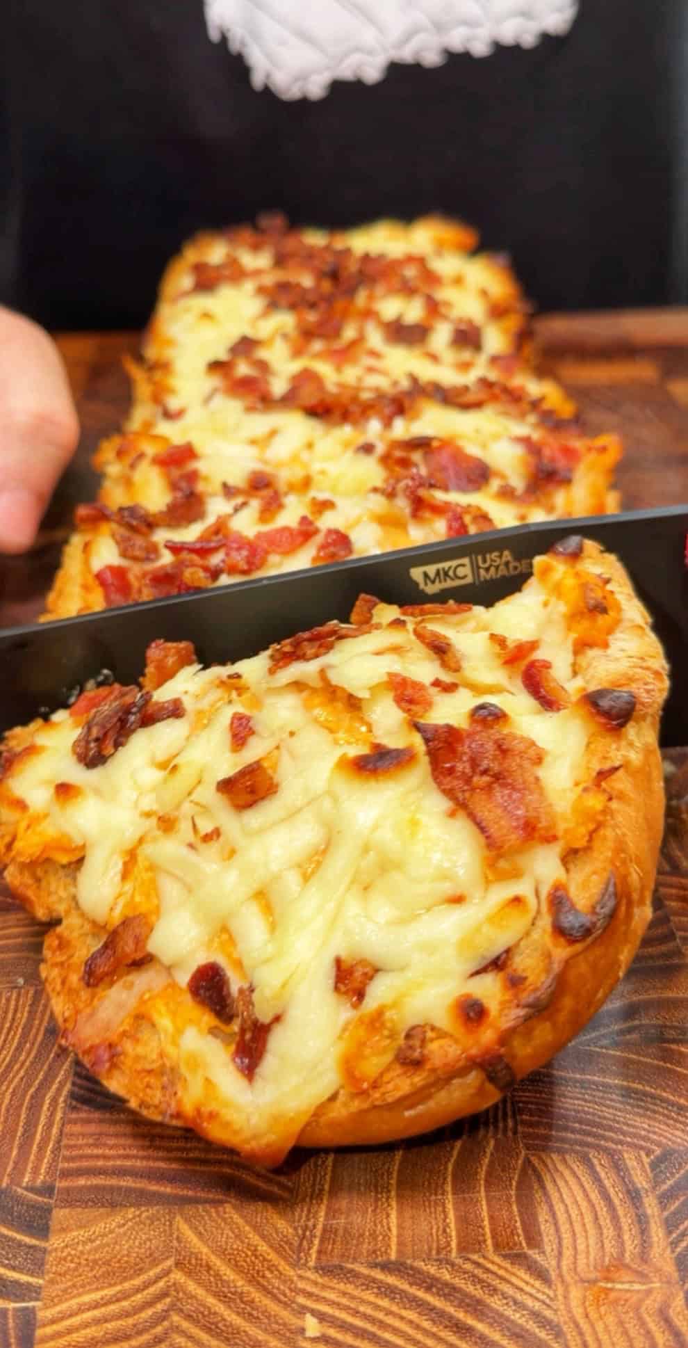 A person slices cheesy bread topped with bacon on a wooden cutting board. The bread looks golden and crisp with melted cheese and bacon bits on top.