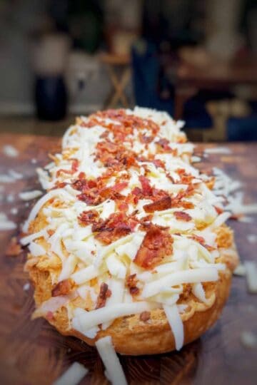 A close-up of a loaf of bread topped with shredded cheese and crispy bacon pieces, resting on a wooden surface with a blurred background.