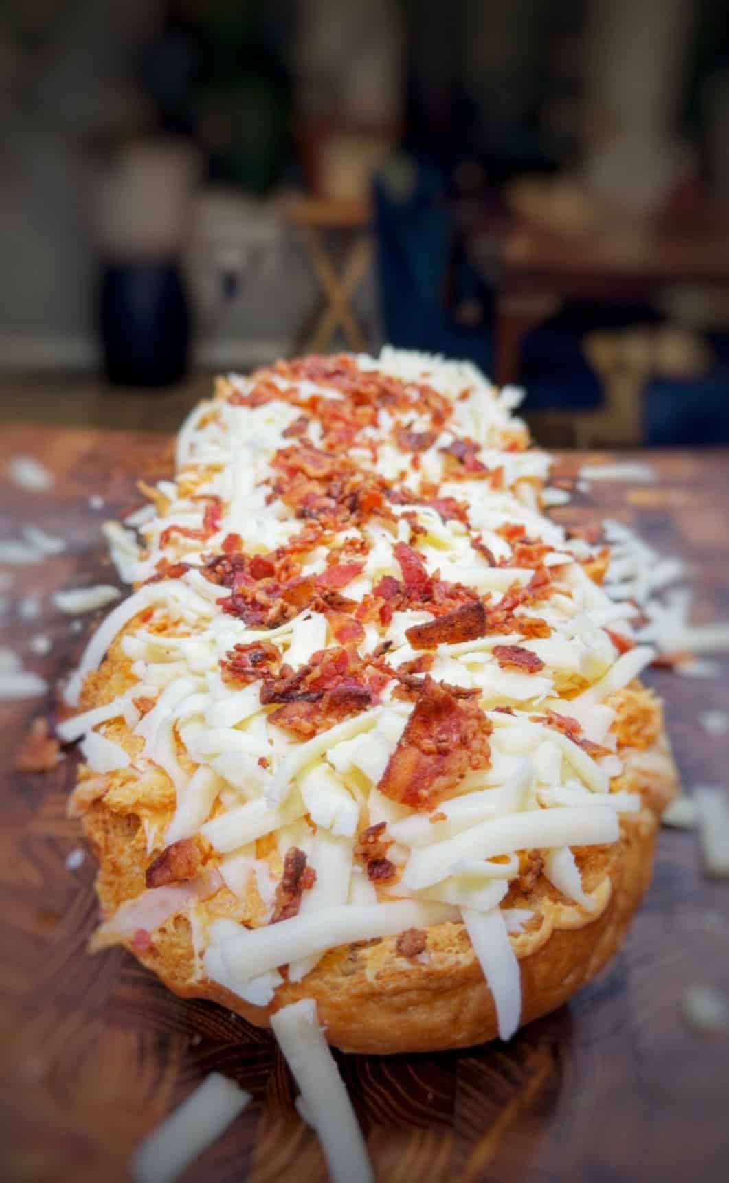 A close-up of a loaf of bread topped with shredded cheese and crispy bacon pieces, resting on a wooden surface with a blurred background.