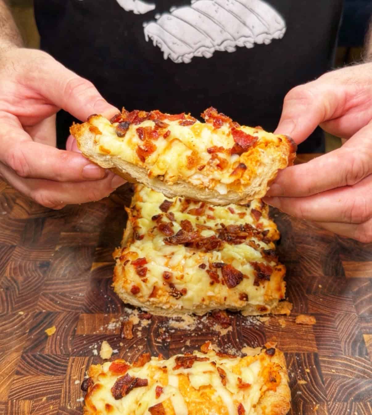 A person holds a cheesy bread slice topped with bacon bits above a wooden board, with more similar cheesy bread slices stacked below. The person is wearing a black shirt with a white graphic.