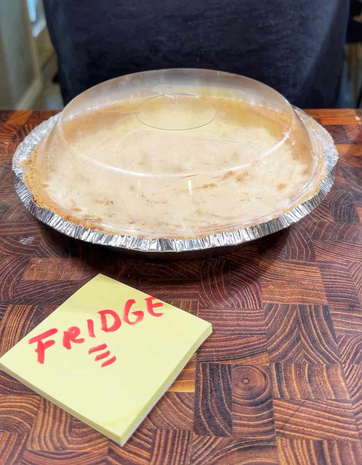 A covered aluminum pie tin sits on a wooden surface with a yellow sticky note in front labeled FRIDGE in red marker.