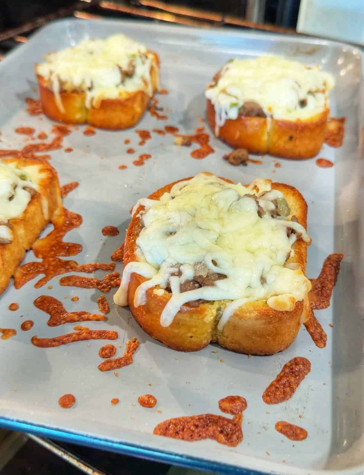 Slices of toasted bread topped with ground meat and melted mozzarella cheese on a baking sheet, with bits of melted cheese scattered around.