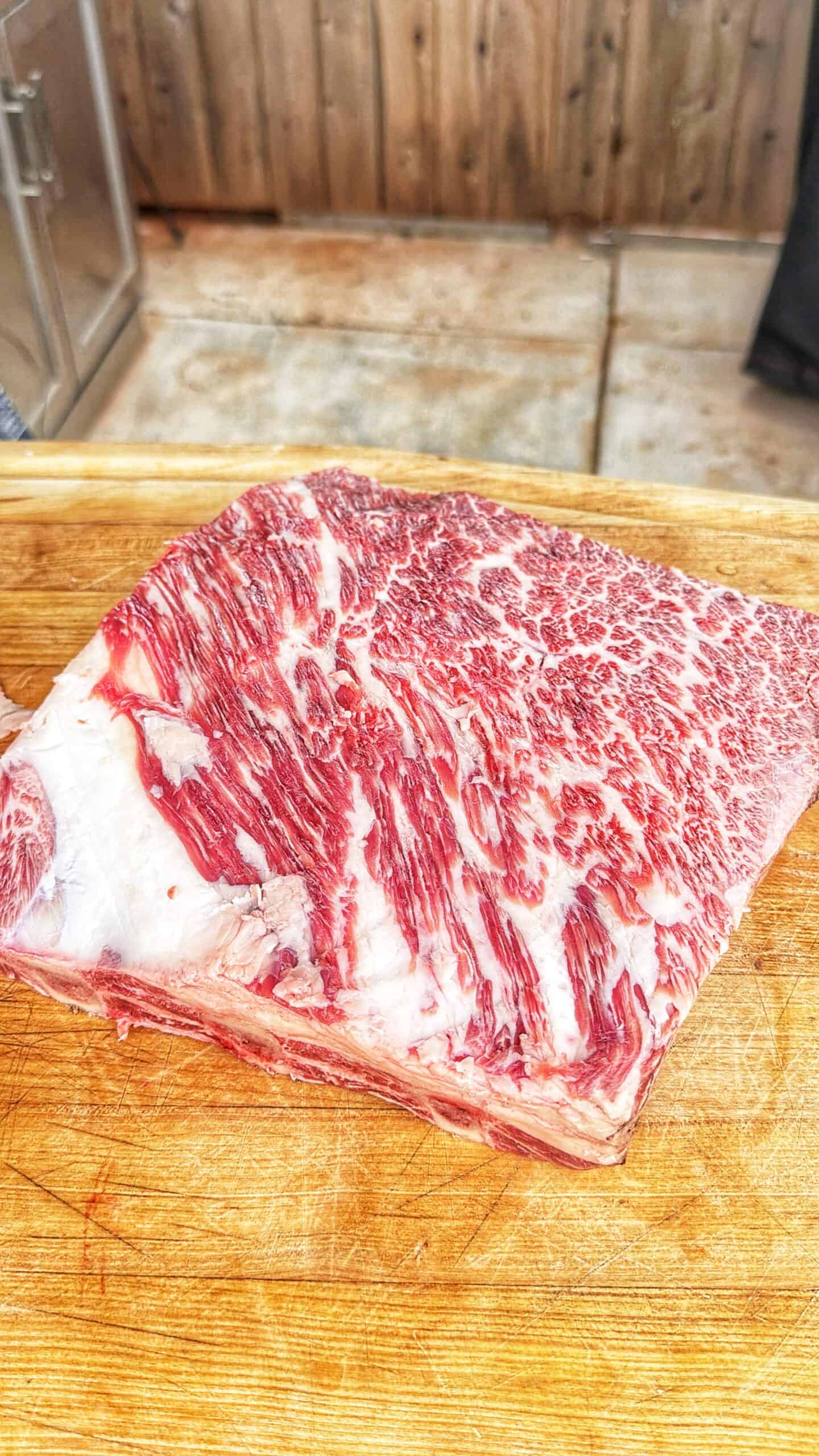 A thick, rectangular cut of marbled raw beef sits on a wooden cutting board. The beef displays prominent white fat marbling throughout the red meat. A rustic wooden wall is in the background.