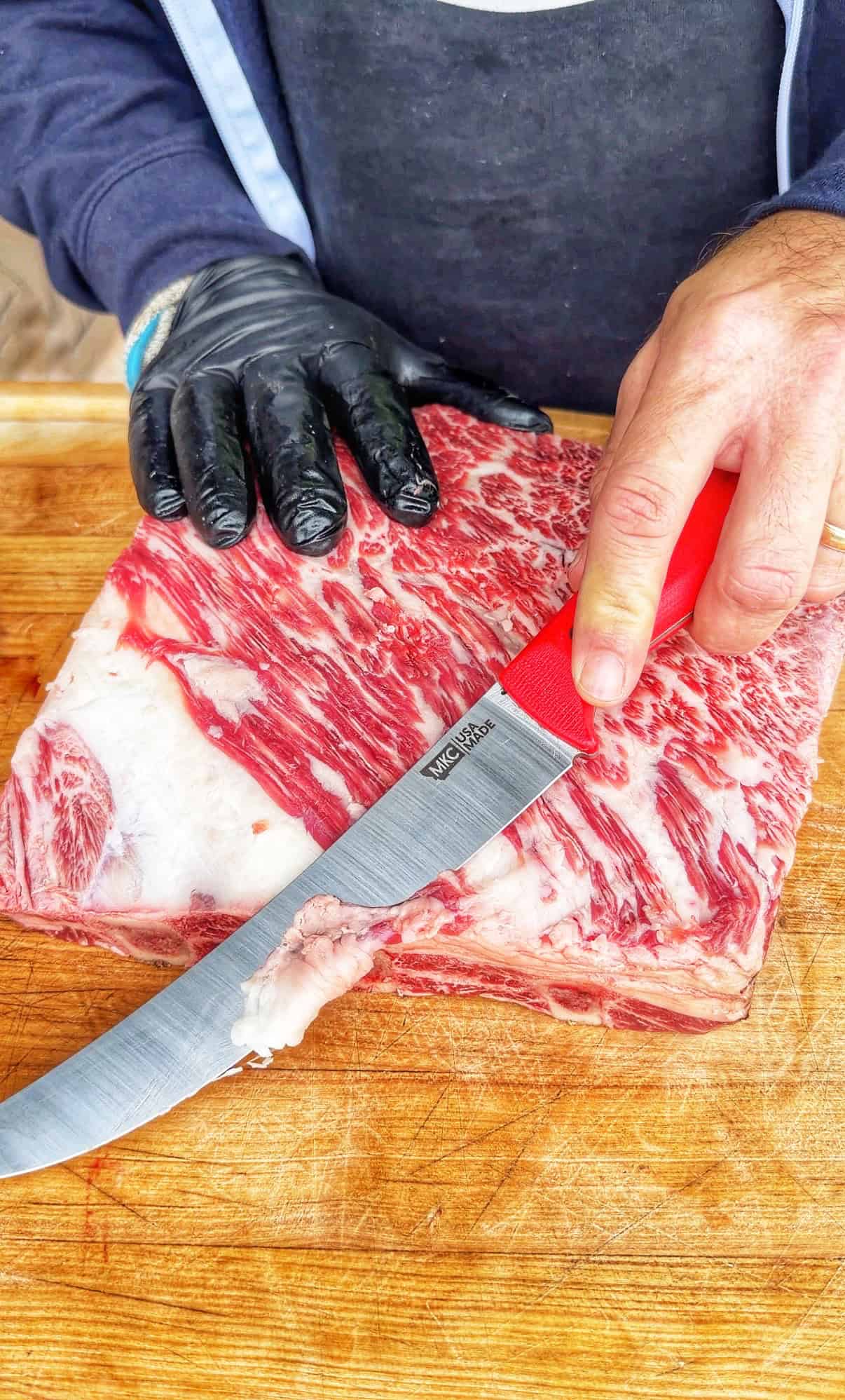 A person wearing a black glove on one hand holds a large cut of marbled raw beef on a wooden board, while the other hand uses a red-handled knife to trim fat from the meat.