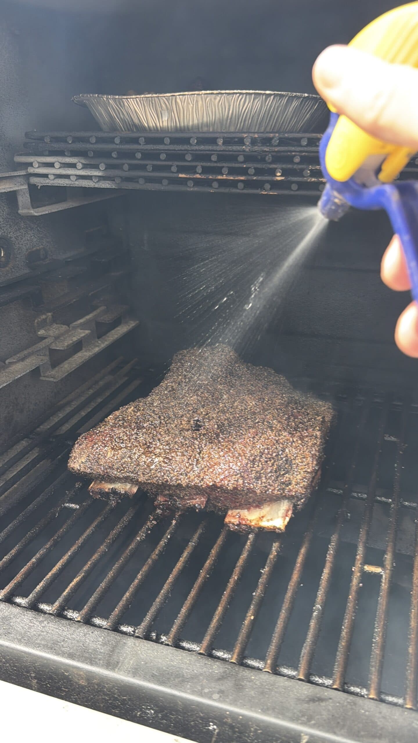 A person sprays liquid onto a large piece of seasoned meat smoking on a grill rack inside a smoker, with a foil pan on an upper rack.
