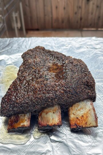 A large, cooked beef rib with a dark, spiced crust rests on a sheet of aluminum foil, exposing three cleaned rib bones. The background shows a wooden fence and an outdoor kitchen area.