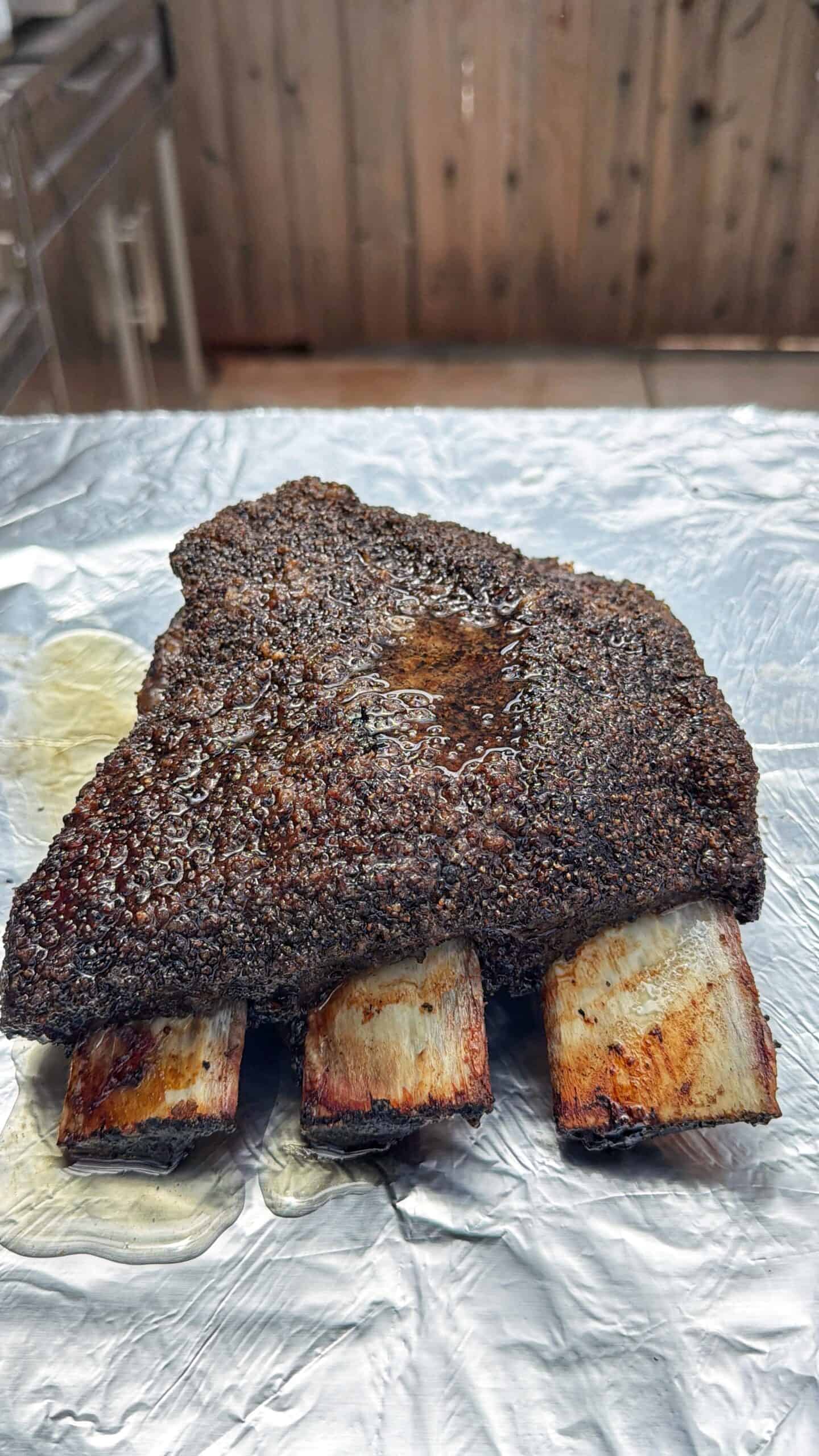 A large, cooked beef rib with a dark, spiced crust rests on a sheet of aluminum foil, exposing three cleaned rib bones. The background shows a wooden fence and an outdoor kitchen area.