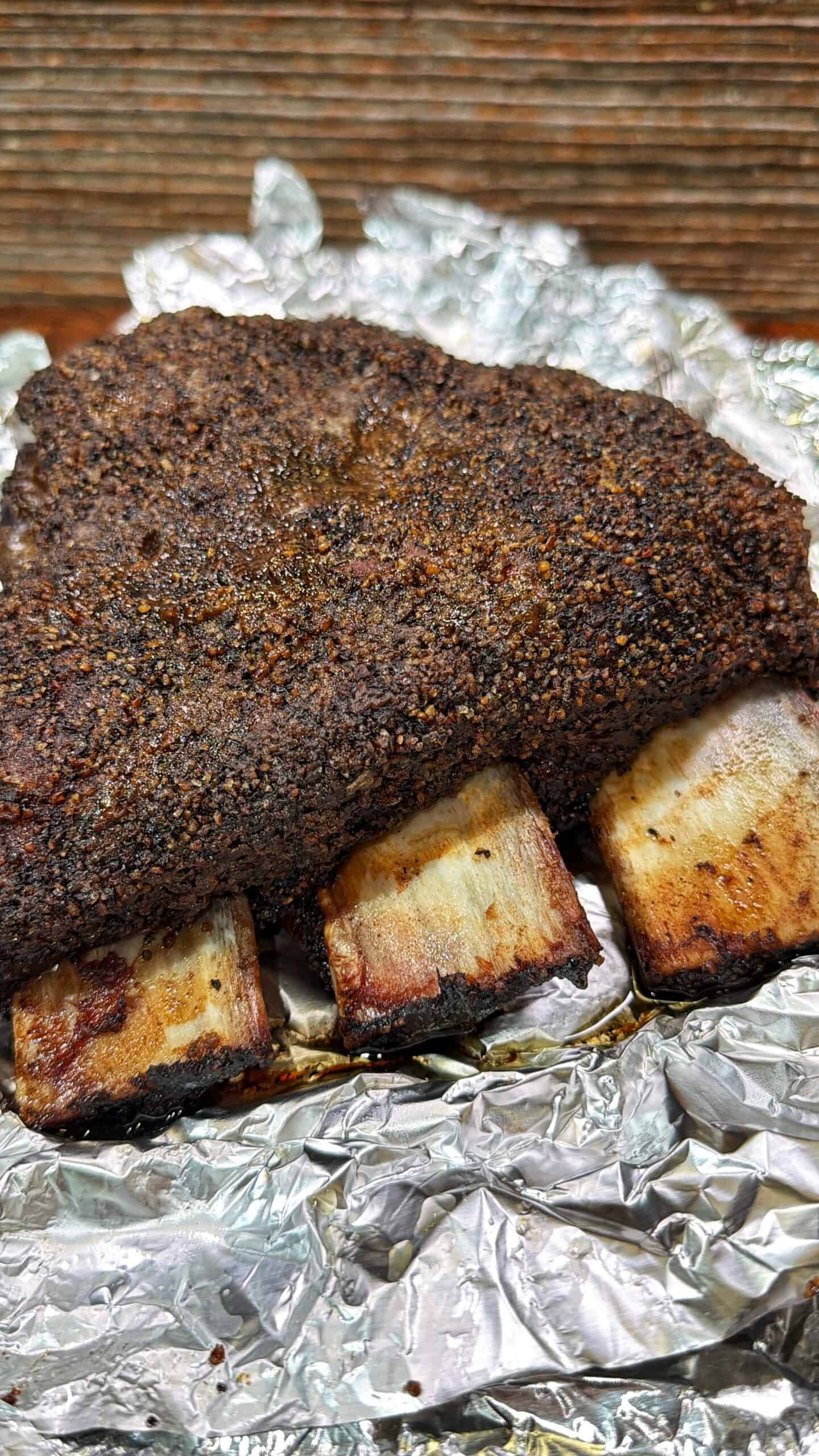 A rack of smoked beef ribs with a dark, seasoned bark rests on crumpled aluminum foil against a wooden background.