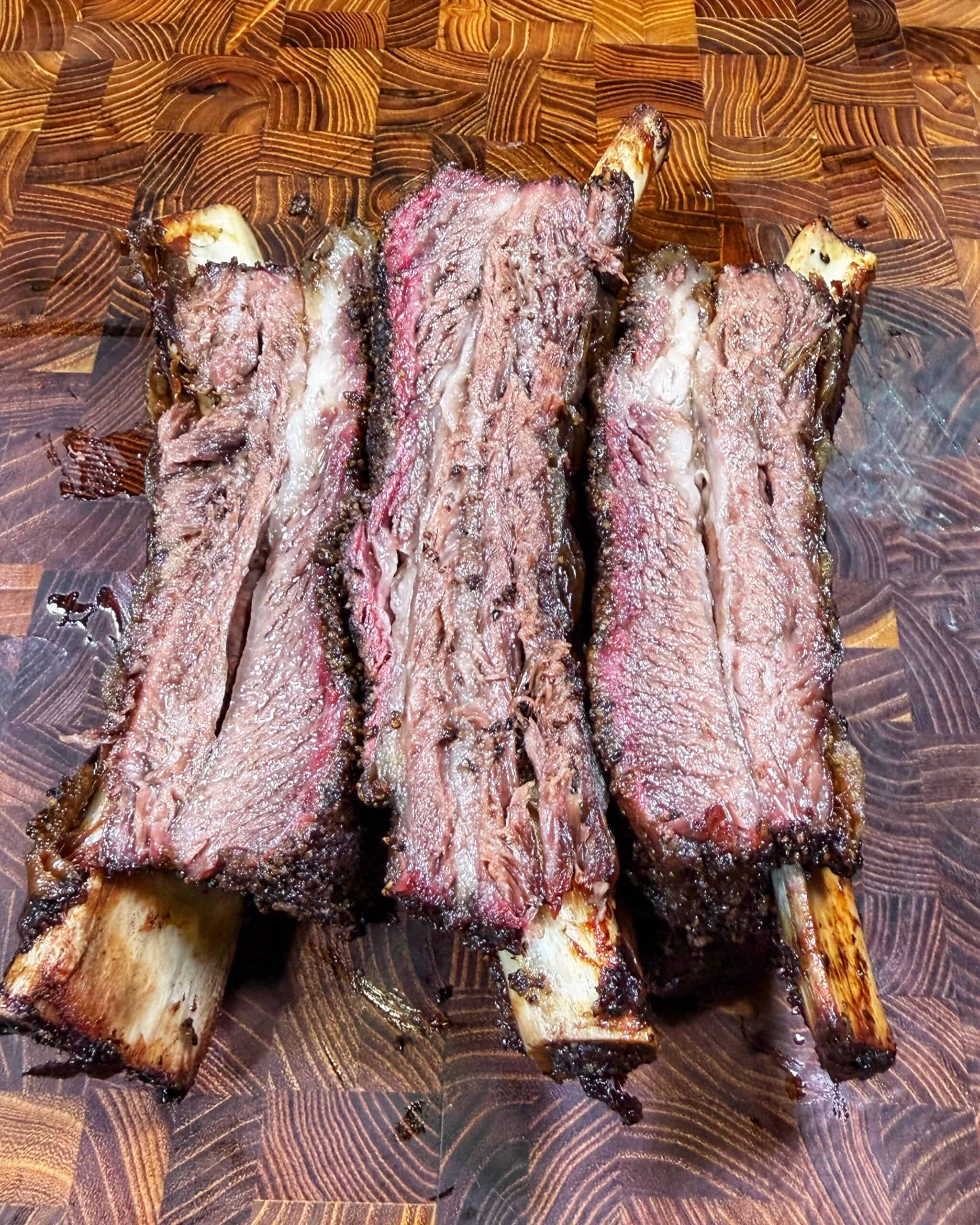 Three large, smoked beef ribs cut in half, revealing a juicy, pink interior and a dark, seasoned crust, are arranged on a wooden cutting board with a rich, geometric pattern.