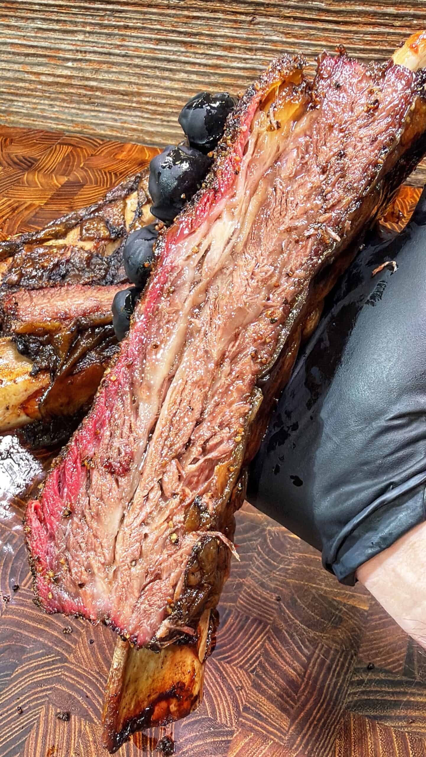 A gloved hand holds a large, juicy, smoked beef rib with a visible smoke ring, tender meat, and a bone, set against a wooden background.