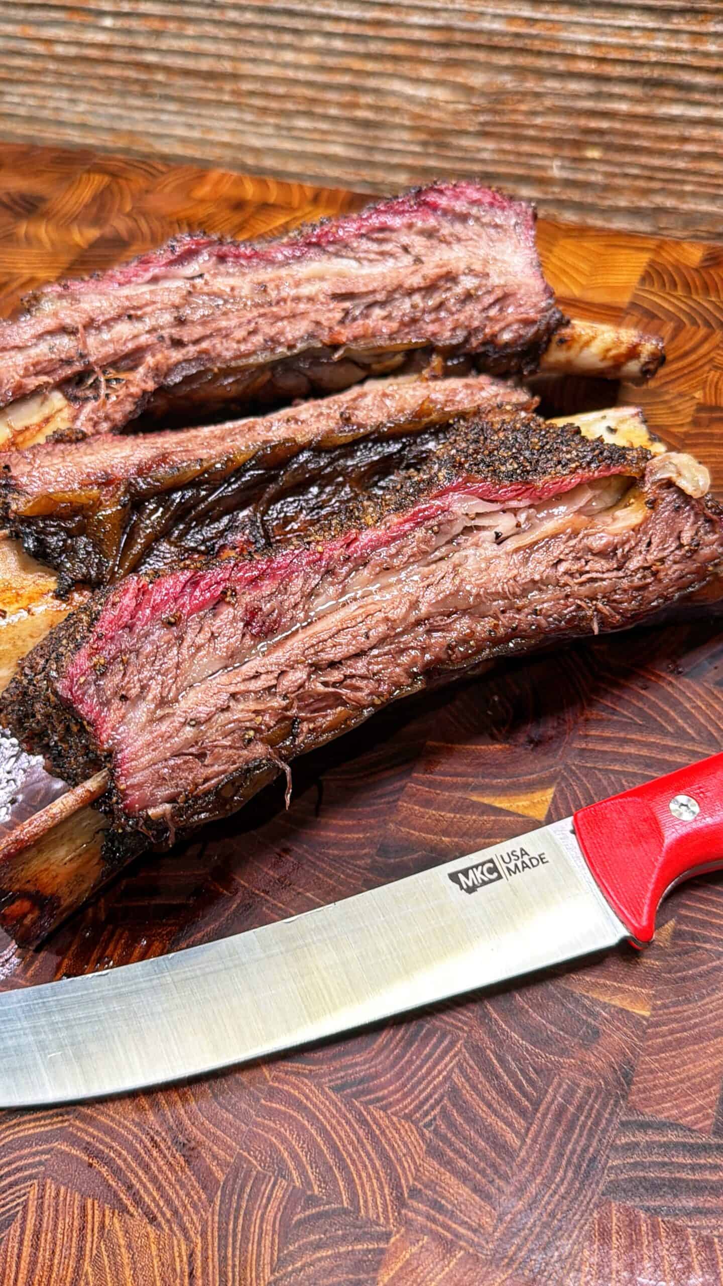 Three thick, juicy smoked beef ribs rest on a wooden cutting board next to a sharp knife with a red handle and a visible USA Made logo on the blade.