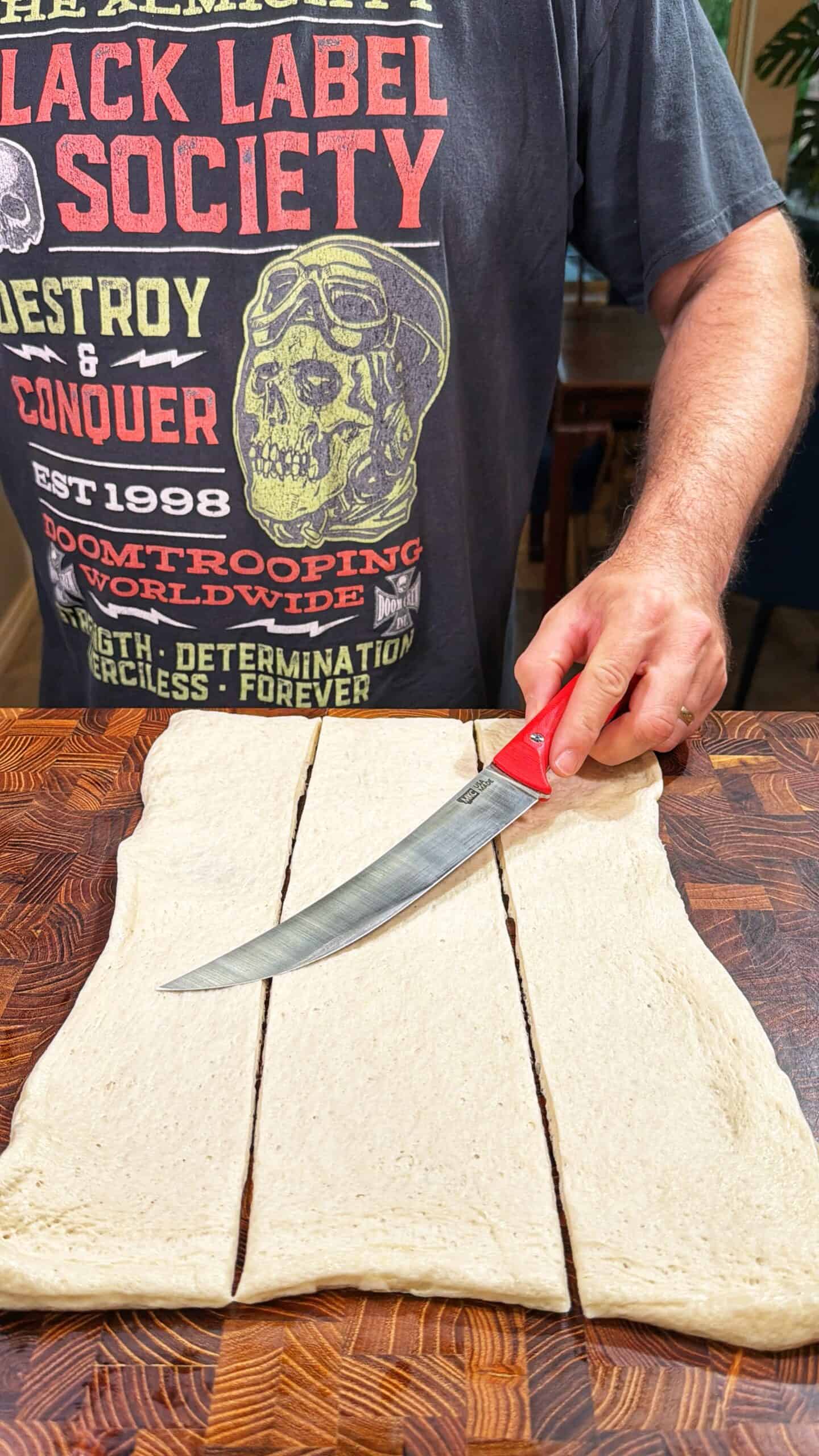 A person in a graphic T-shirt slices dough into three strips on a wooden cutting board using a large knife with a red handle.