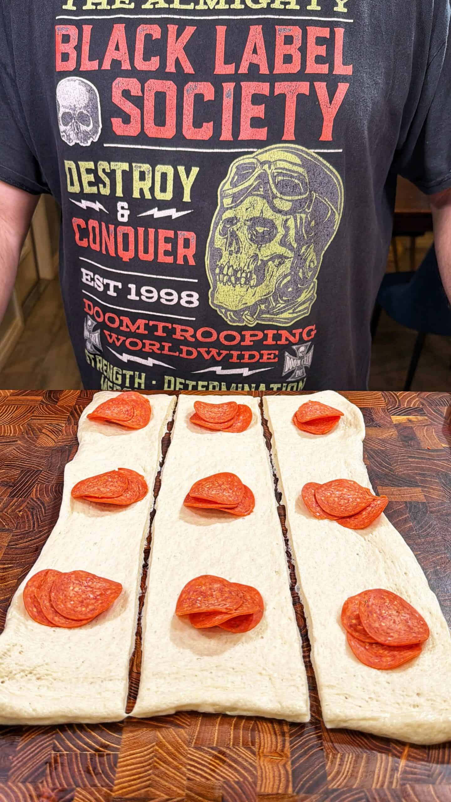 A person wearing a Black Label Society T-shirt stands behind a wooden surface with three strips of pizza dough topped with slices of pepperoni.