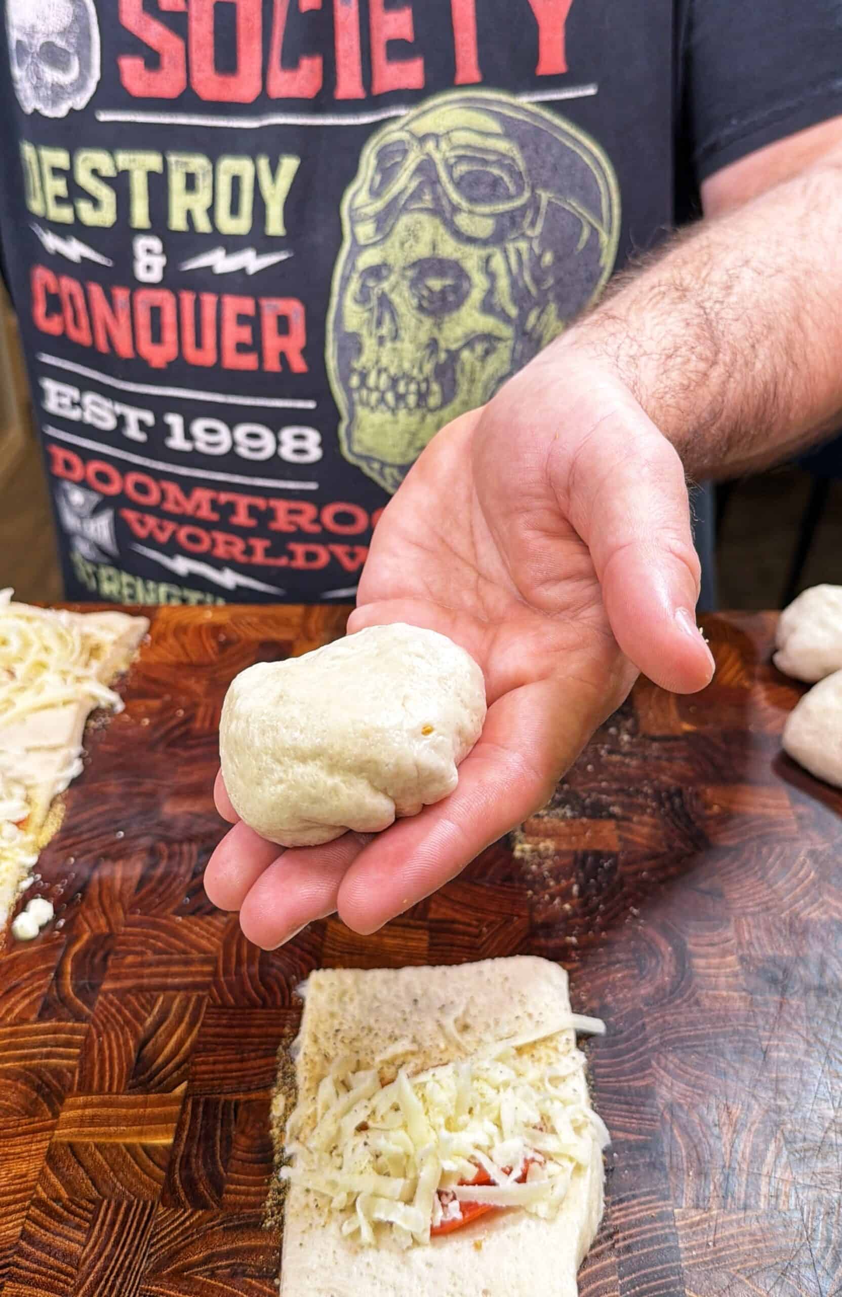 A person holds a ball of dough in their palm over a wooden surface with grated cheese and a rectangular piece of dough. The person wears a black T-shirt with bold, colorful text and a skull graphic.