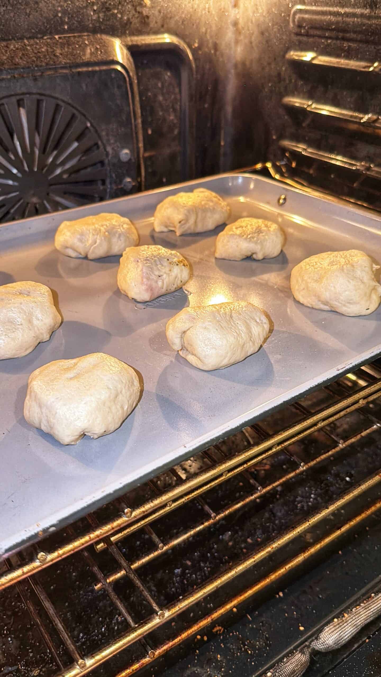 Nine pieces of dough are baking on a large silver baking sheet inside an oven, with the oven door open and the heating element visible in the background.