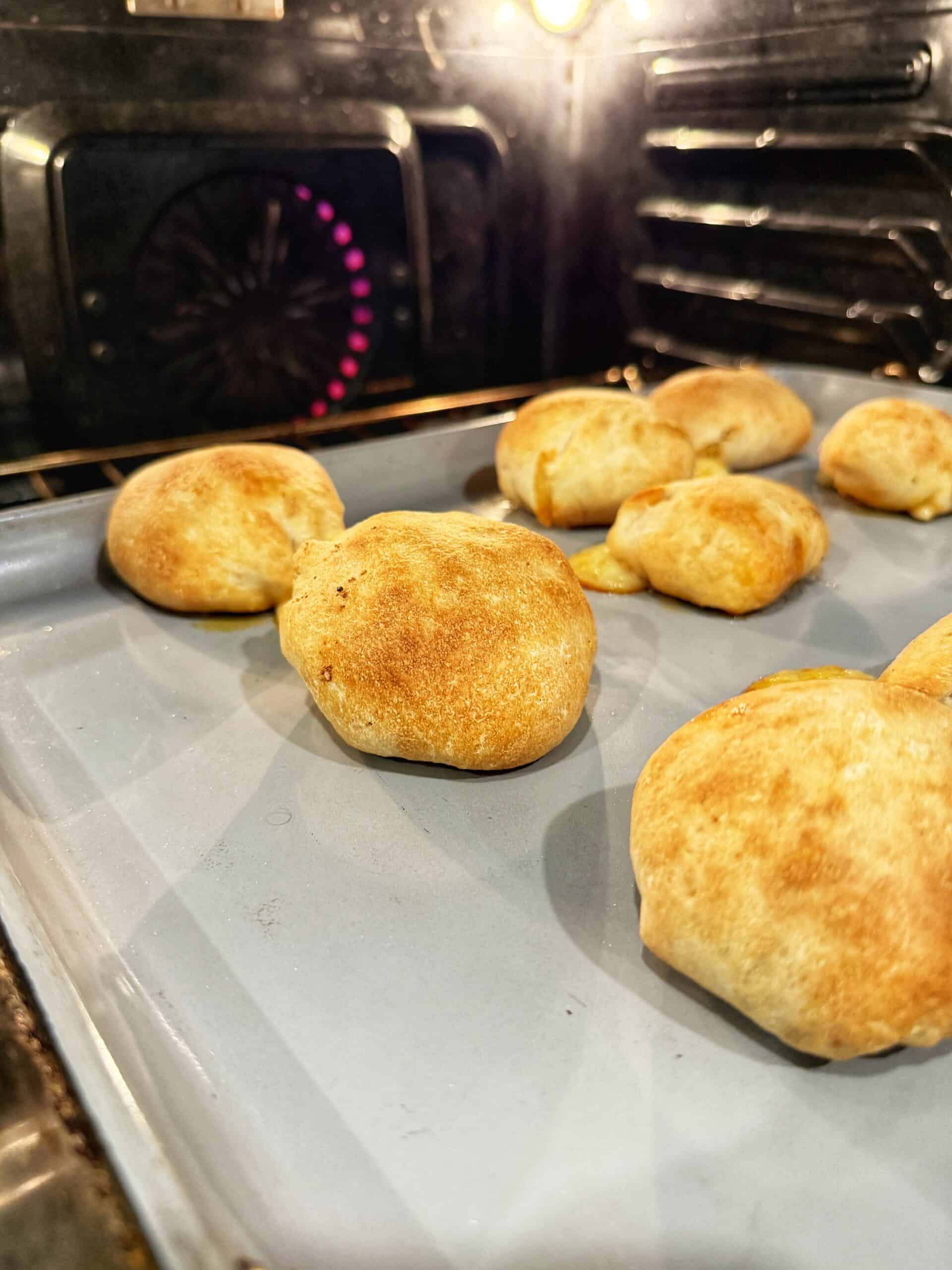 Golden brown rolls baking on a light blue baking sheet inside an oven, with the oven light illuminating the freshly baked bread.