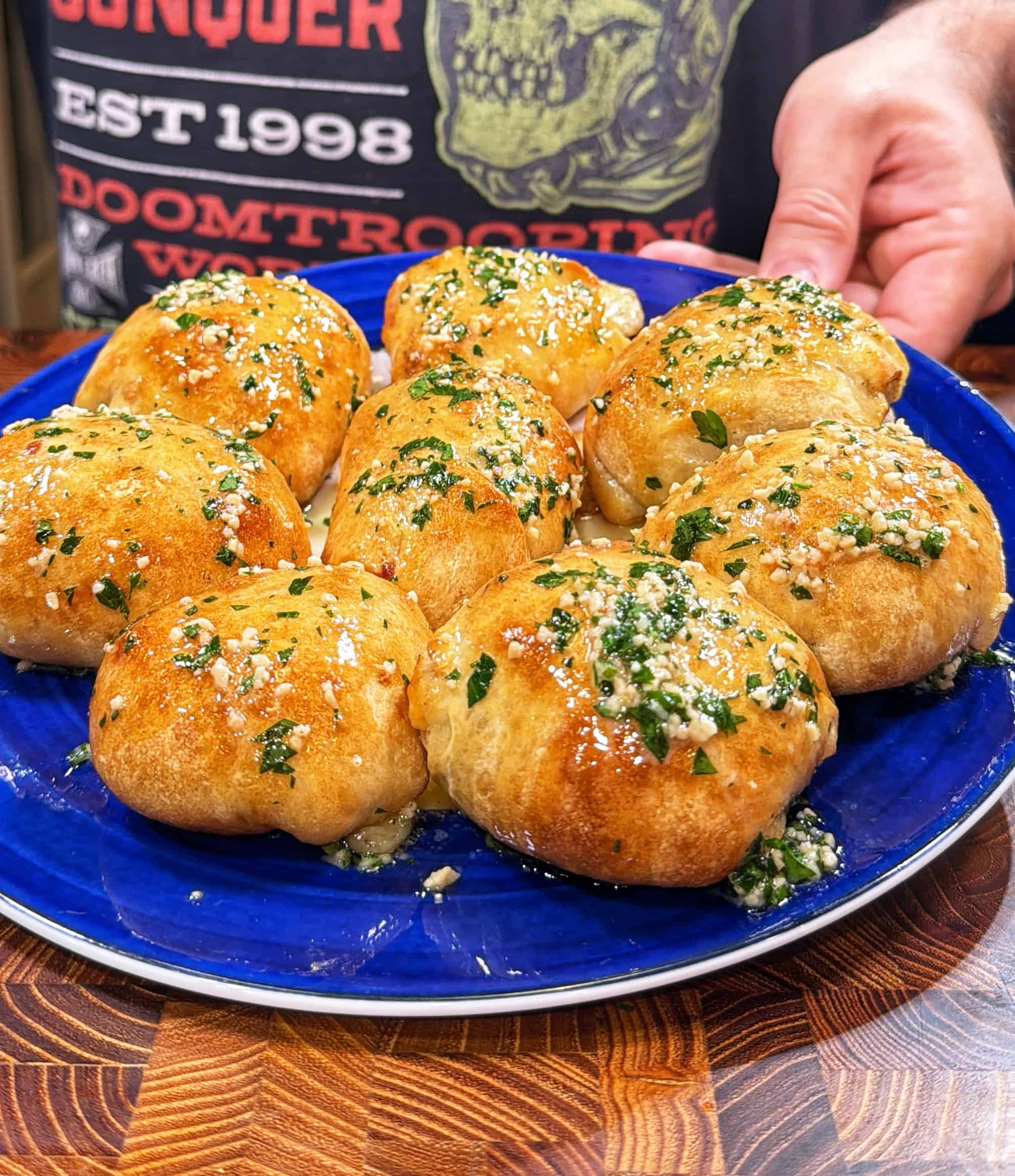 A blue plate with eight golden-brown baked rolls topped with chopped herbs and grated cheese. A person wearing a graphic t-shirt is holding the plate.