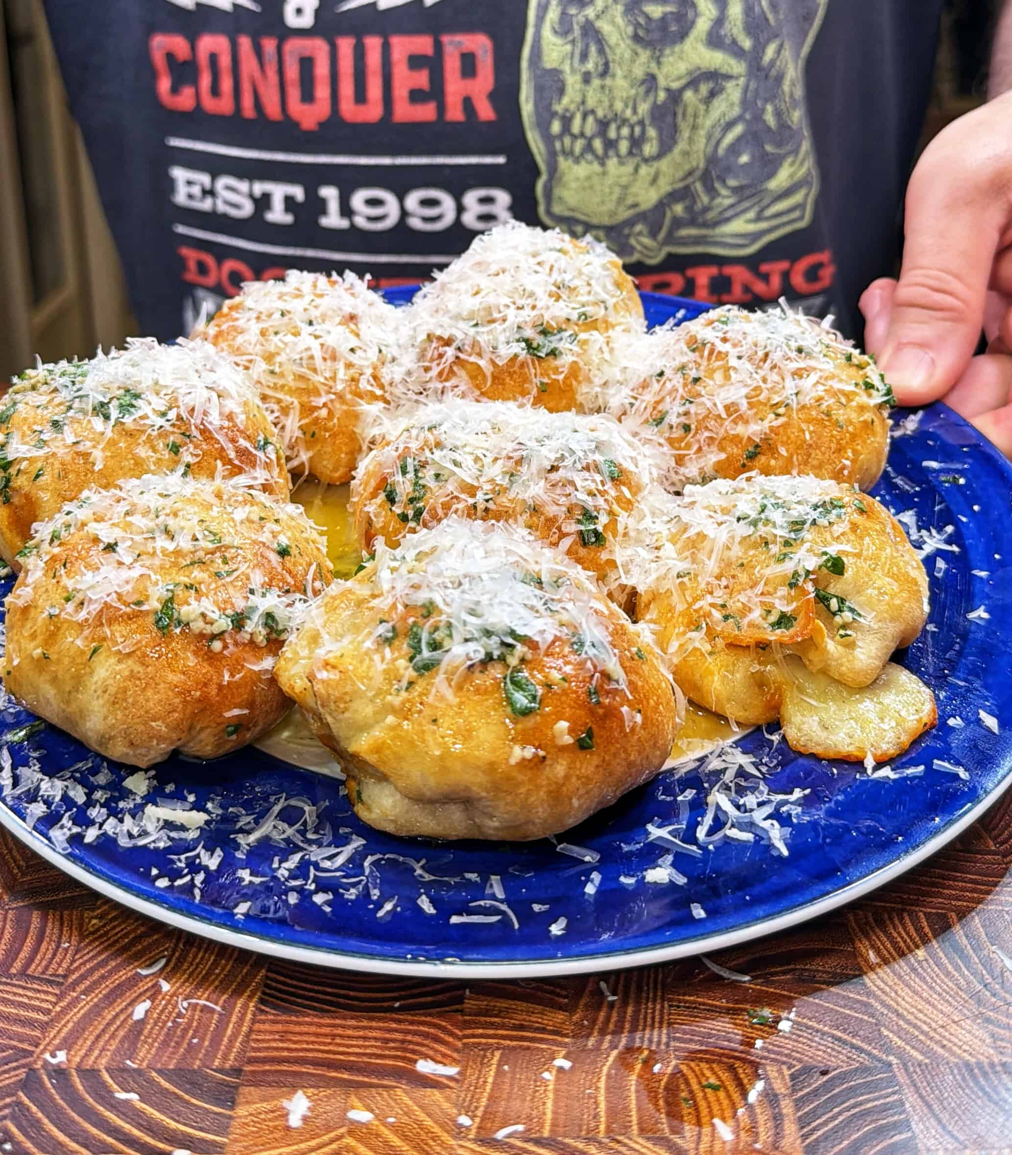 A blue plate filled with golden, baked garlic knots topped with grated cheese and herbs. In the background, a person wearing a dark shirt with graphic text and a skull image holds the plate.