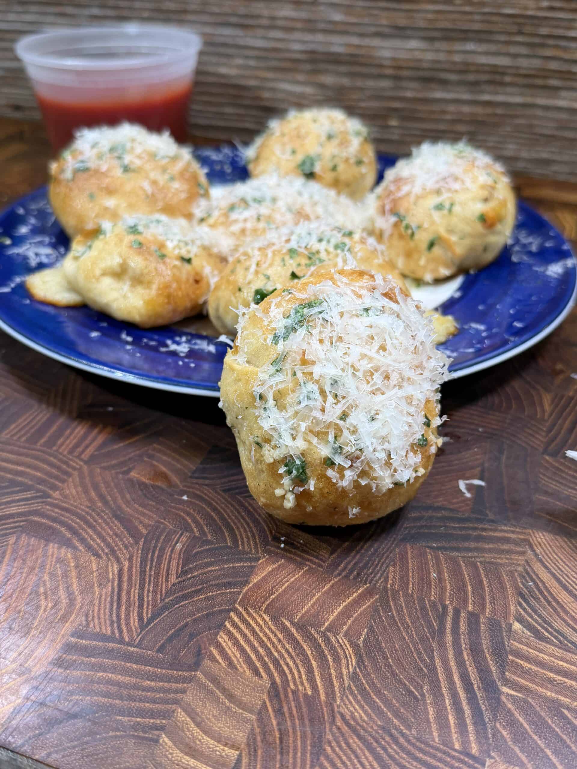 A close-up of a plate with several golden-brown garlic knots topped with shredded cheese and herbs. One knot is in the foreground, and a small cup of marinara sauce is in the background.