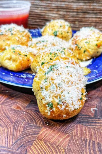 A close-up of cheesy garlic bread bites topped with grated parmesan and herbs on a blue plate, with a small container of marinara sauce in the background on a wooden surface.