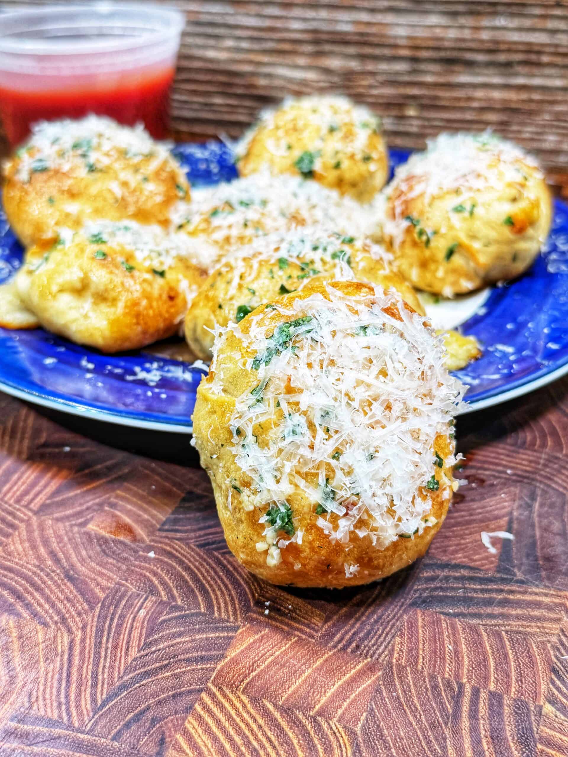 A close-up of cheesy garlic bread bites topped with grated parmesan and herbs on a blue plate, with a small container of marinara sauce in the background on a wooden surface.