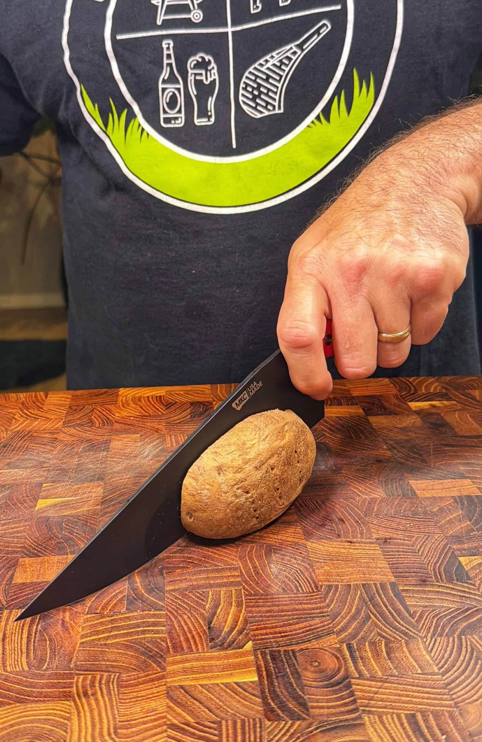 A person wearing a dark T-shirt is slicing a brown potato on a wooden cutting board with a black knife. The persons left hand holds the potato steady, and they are wearing a gold ring.
