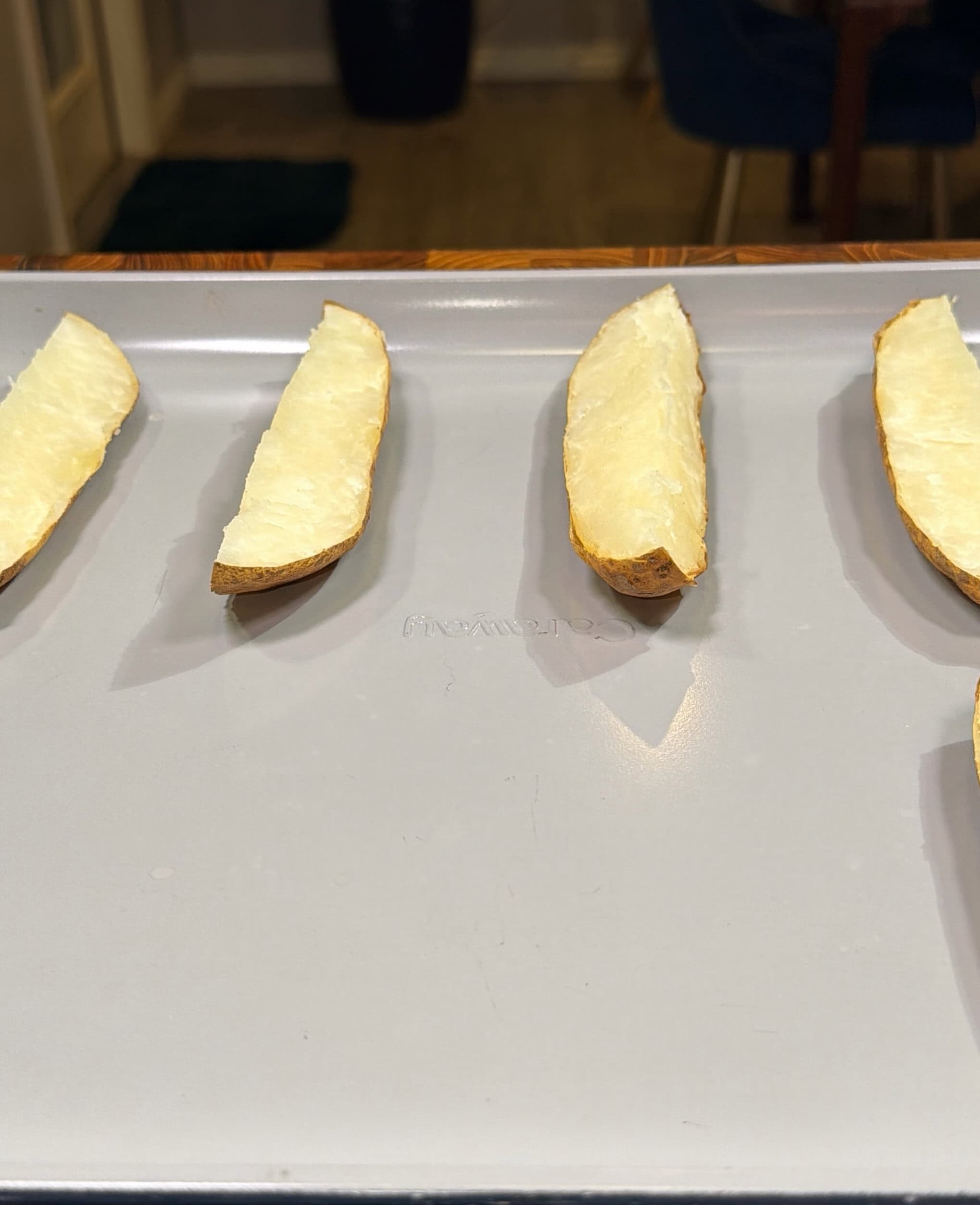 Four potato halves with most of the insides scooped out are arranged in a row on a baking sheet, ready for the next step in preparation, likely to be made into potato skins.