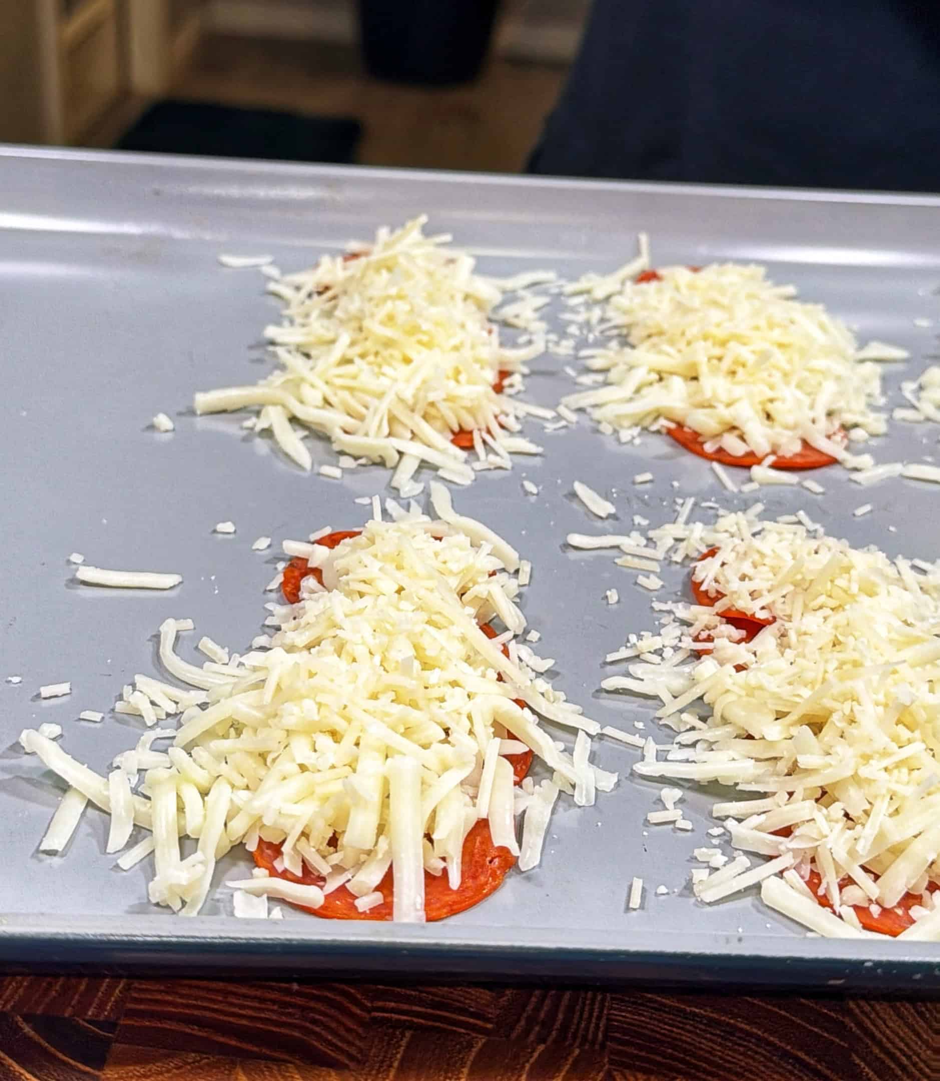 Four small piles of shredded cheese on top of round slices of tomato are arranged on a metal baking sheet, ready to be cooked.