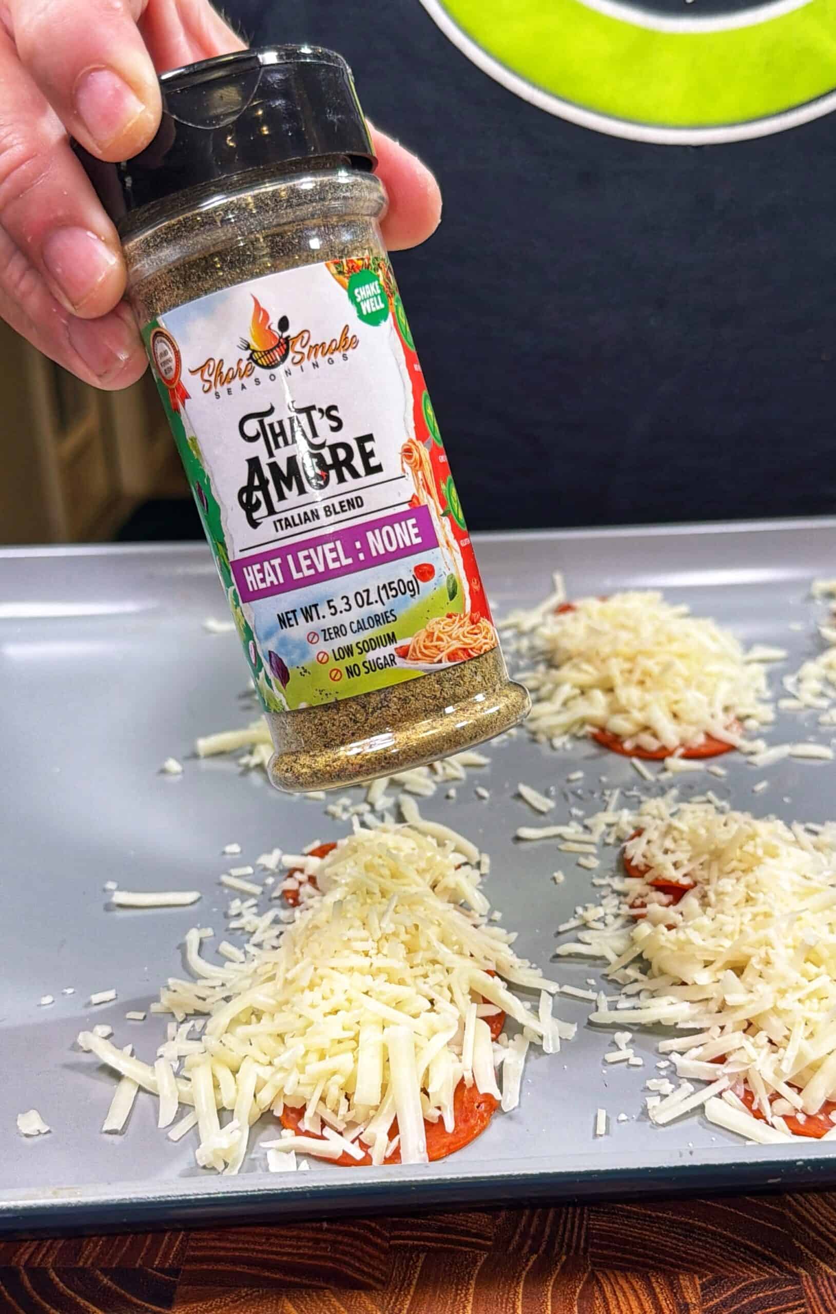 A hand sprinkles Thirs Amore Italian Blend seasoning over mini pizzas topped with shredded cheese on a baking sheet.