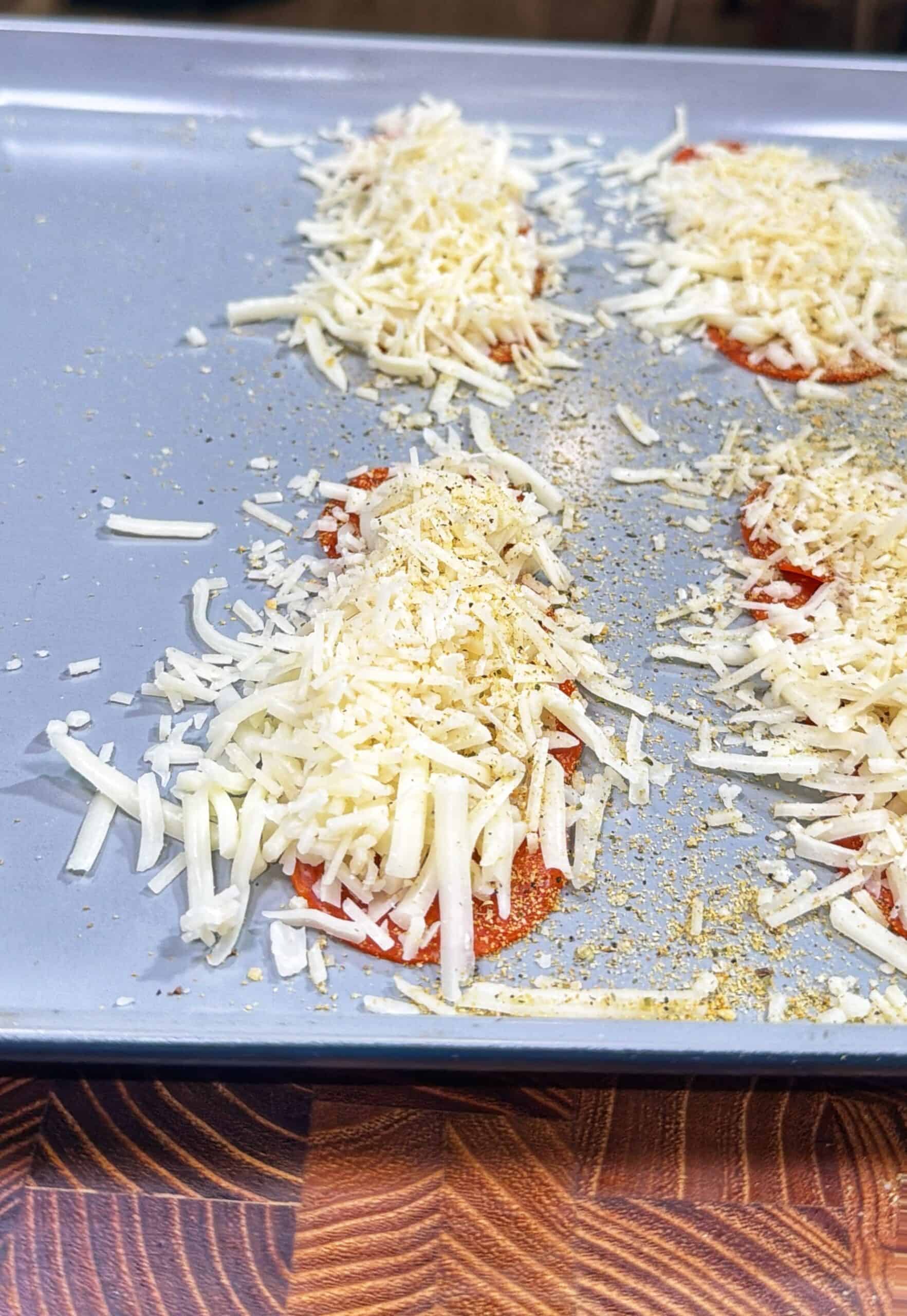 Four rounds of sliced tomato topped with shredded cheese and seasoning are arranged on a gray baking sheet, ready to be baked. The image is taken from above, with a wooden surface at the front.