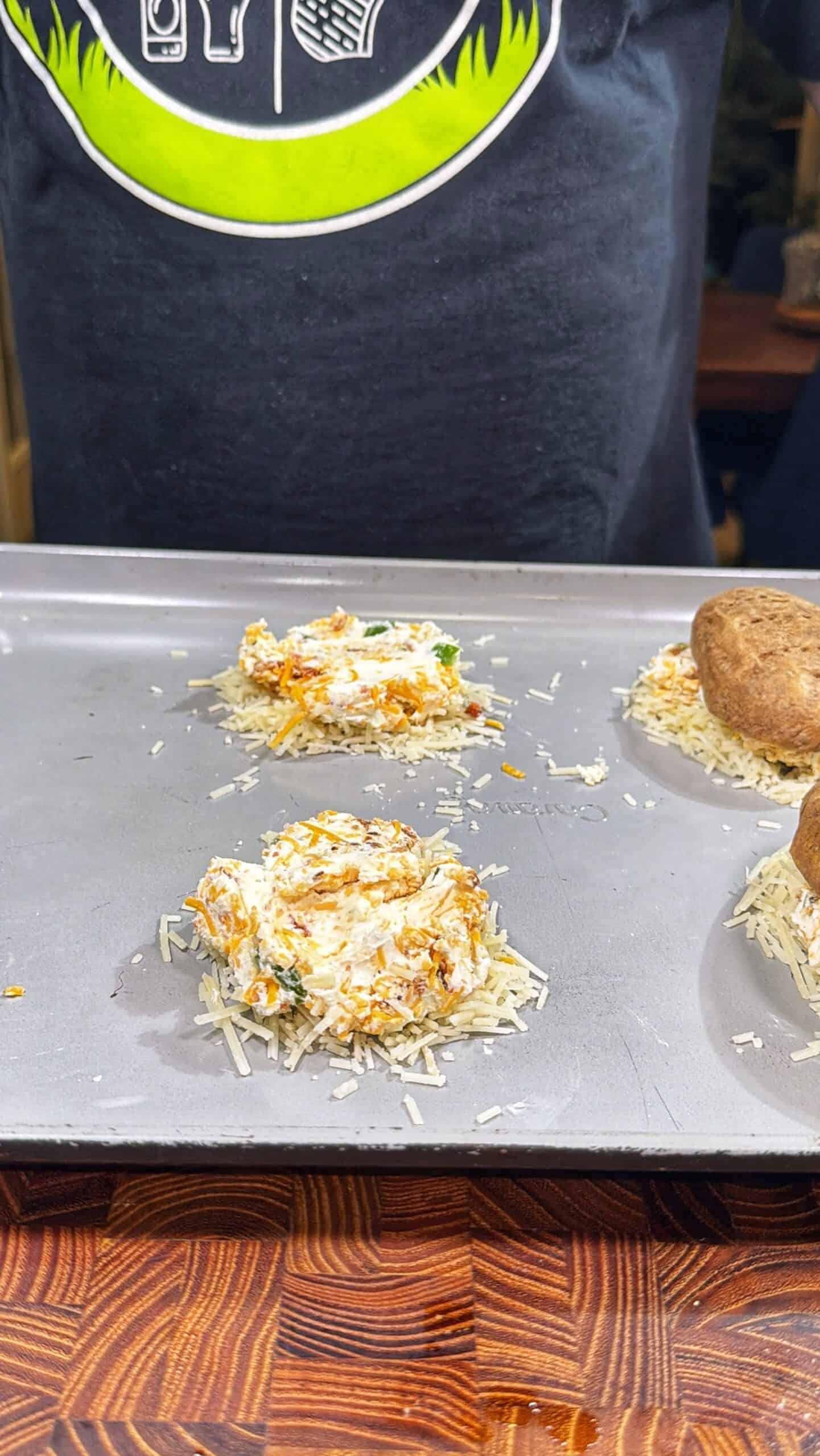 A baking sheet with stuffed mushrooms; two have a filling of shredded cheese and herbs, while another is capped with a whole mushroom. A person in a black shirt with a green logo stands behind the tray.
