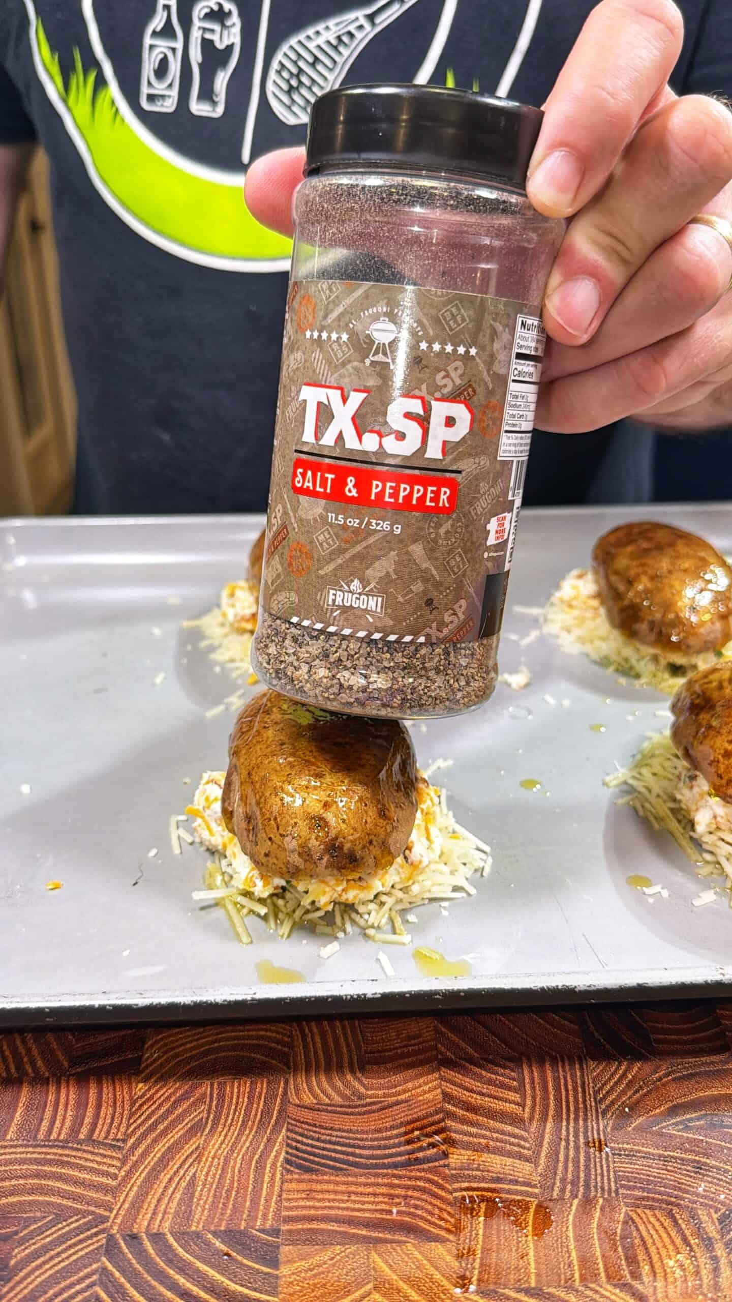 A hand sprinkles TX.SP Salt & Pepper seasoning over a round, seasoned potato on a baking tray with shredded cheese, with a wooden cutting board in the foreground.