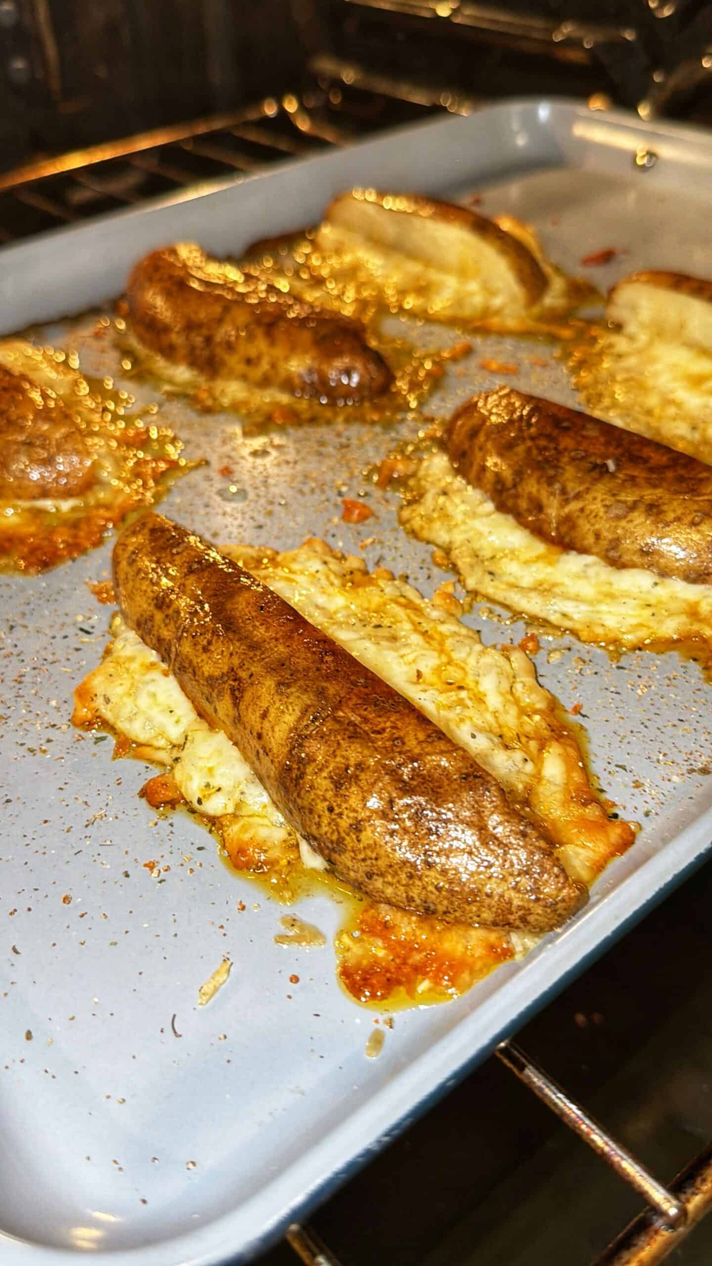 Six browned sausages bake on a gray tray in an oven, with melted cheese oozing out underneath each sausage and pooling on the pan. The sausages appear well-seasoned, and the cheese is bubbling and slightly crisped.
