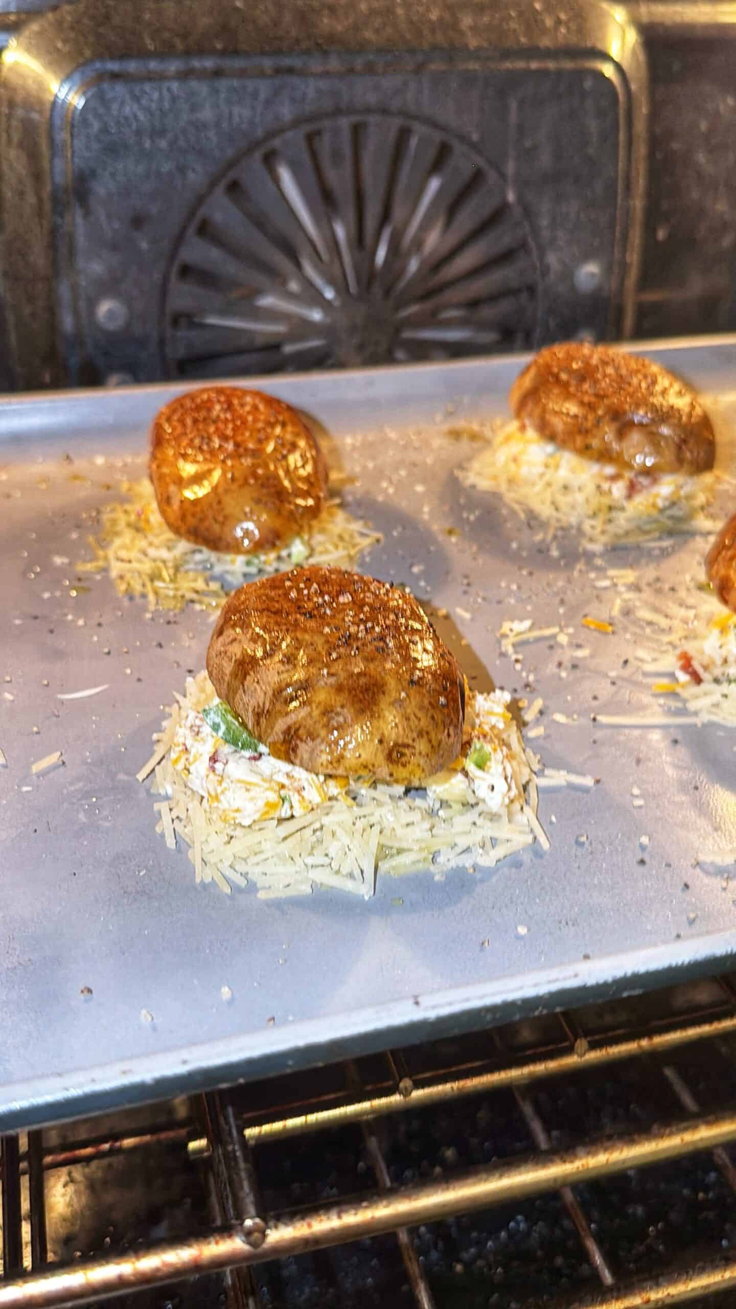 Four stuffed mushrooms sit on a baking sheet in the oven, topped with cheese and seasoning. The oven’s back fan is visible, and the mushrooms are being baked on parchment paper.