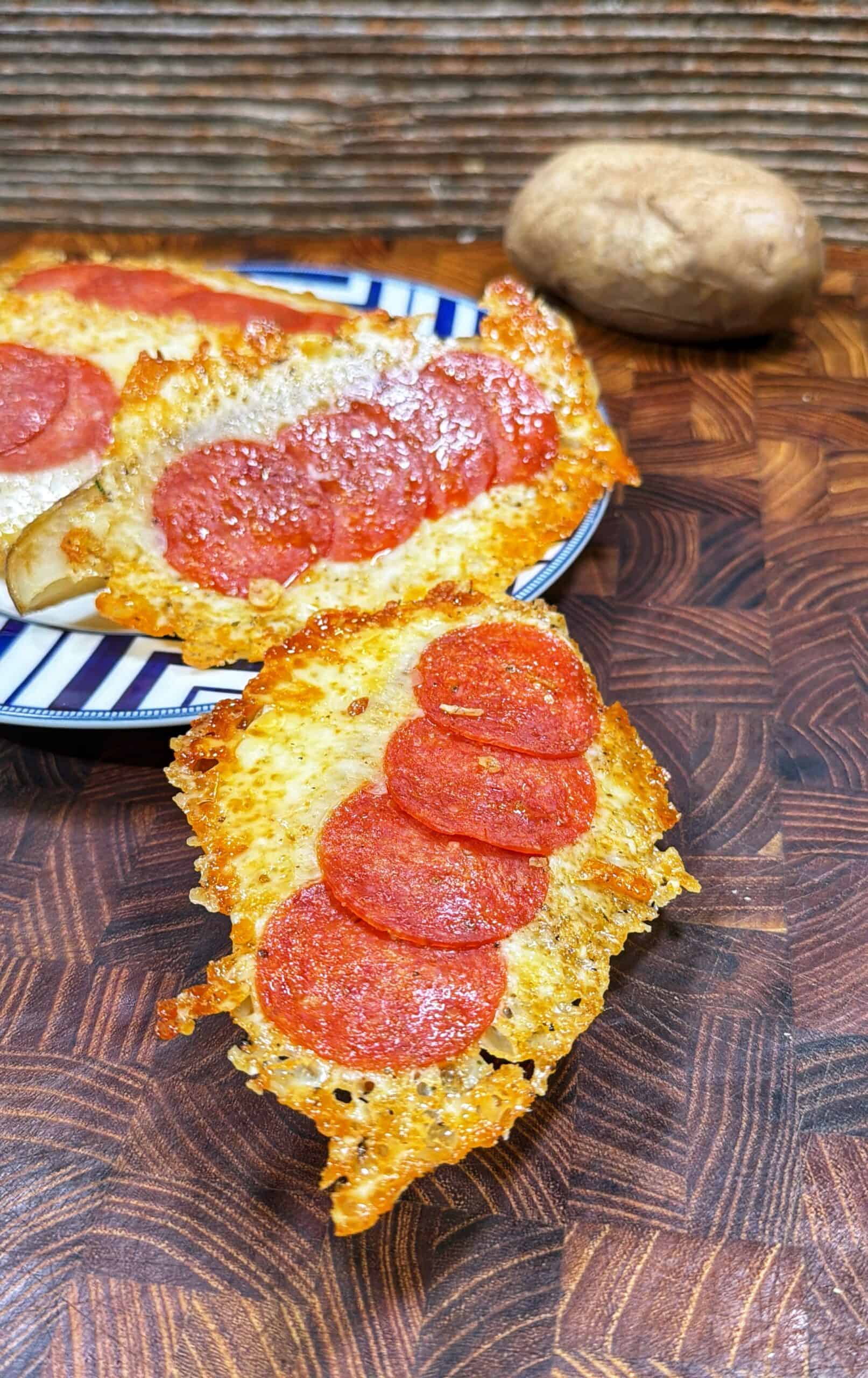 Two potato halves topped with melted cheese and pepperoni slices are displayed on a wooden surface, with one on a striped plate in the background and a whole potato nearby.