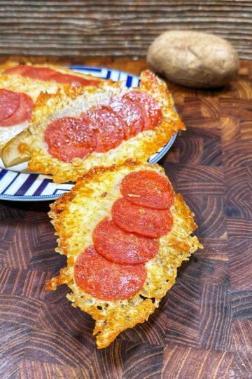Slices of baked potato topped with melted cheese and pepperoni are arranged on a wooden surface, with one piece in the foreground and more pieces on a striped plate in the background. A whole potato sits nearby.