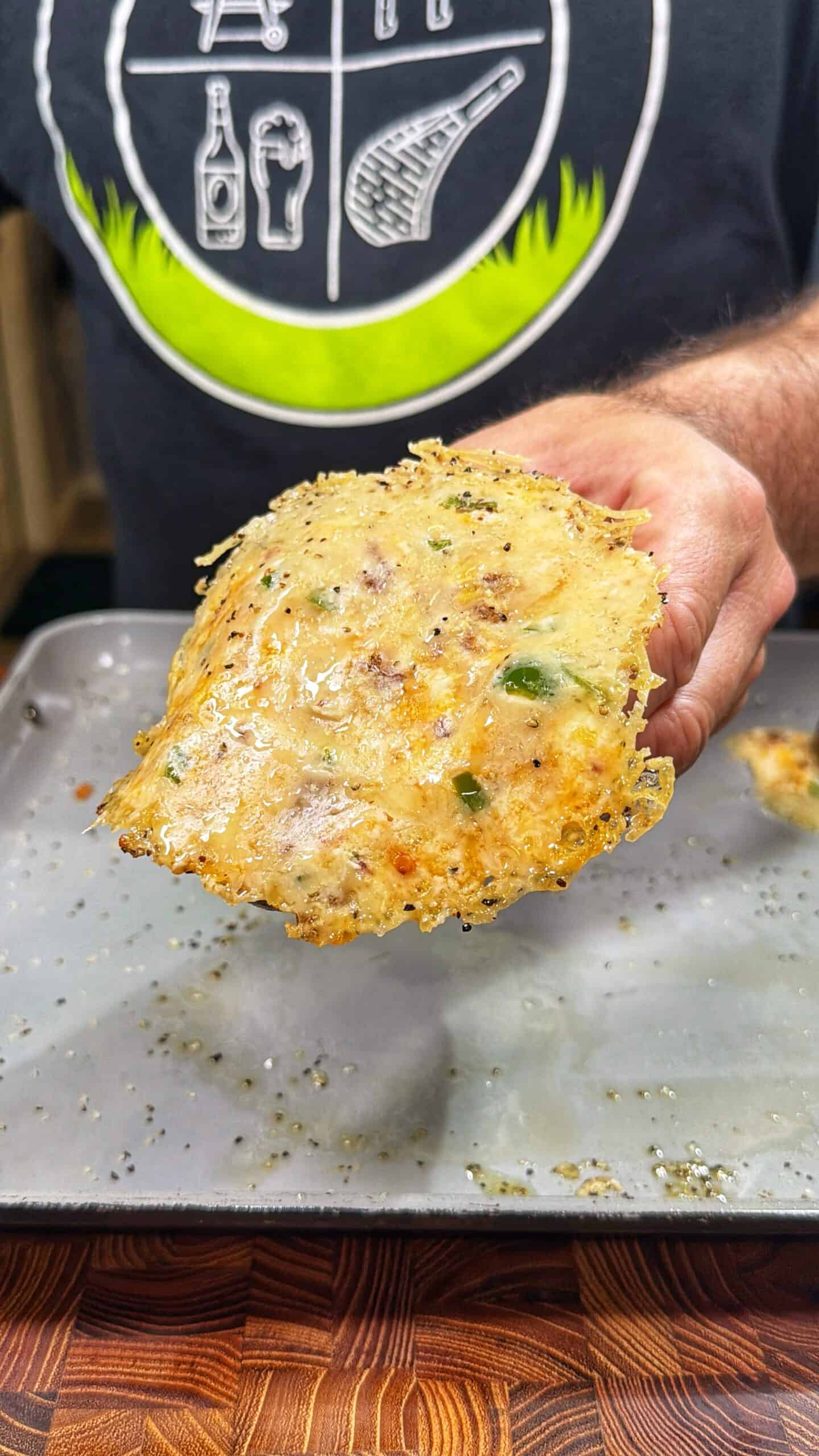 A person holds up a large, crispy cheese crisp with herbs and bits of vegetables over a baking tray. The person is wearing a dark shirt with white graphics and a green stripe.