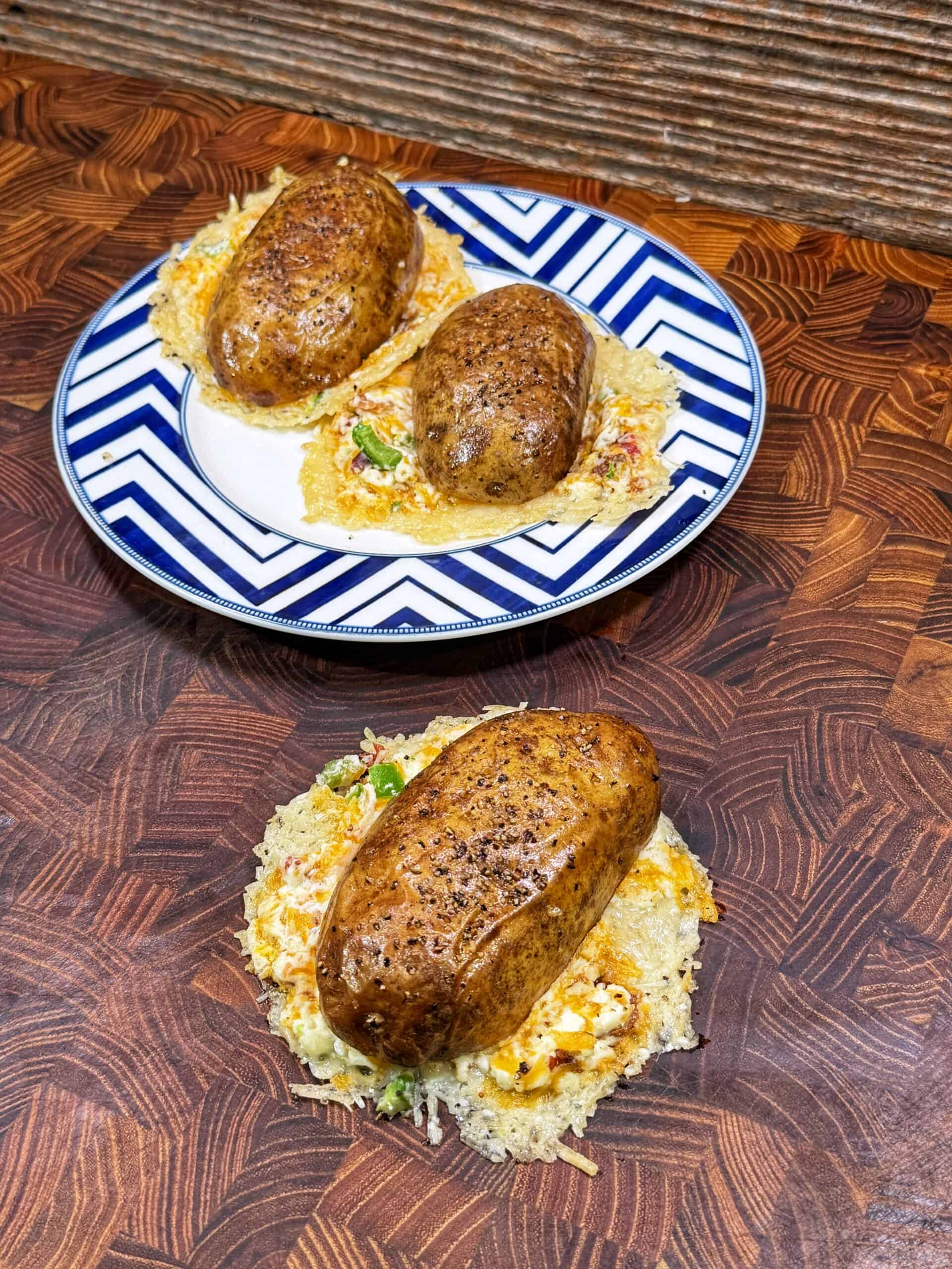 Three baked potatoes rest on crispy cheese nests, topped with sour cream and green onions. Two are on a blue-and-white zigzag plate, while one is directly on a wooden surface with a rich, geometric pattern.