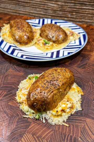 Three baked potatoes topped with melted cheese and herbs, served on a patterned plate and a wooden surface. The potatoes rest on a layer of crispy, golden cheese.