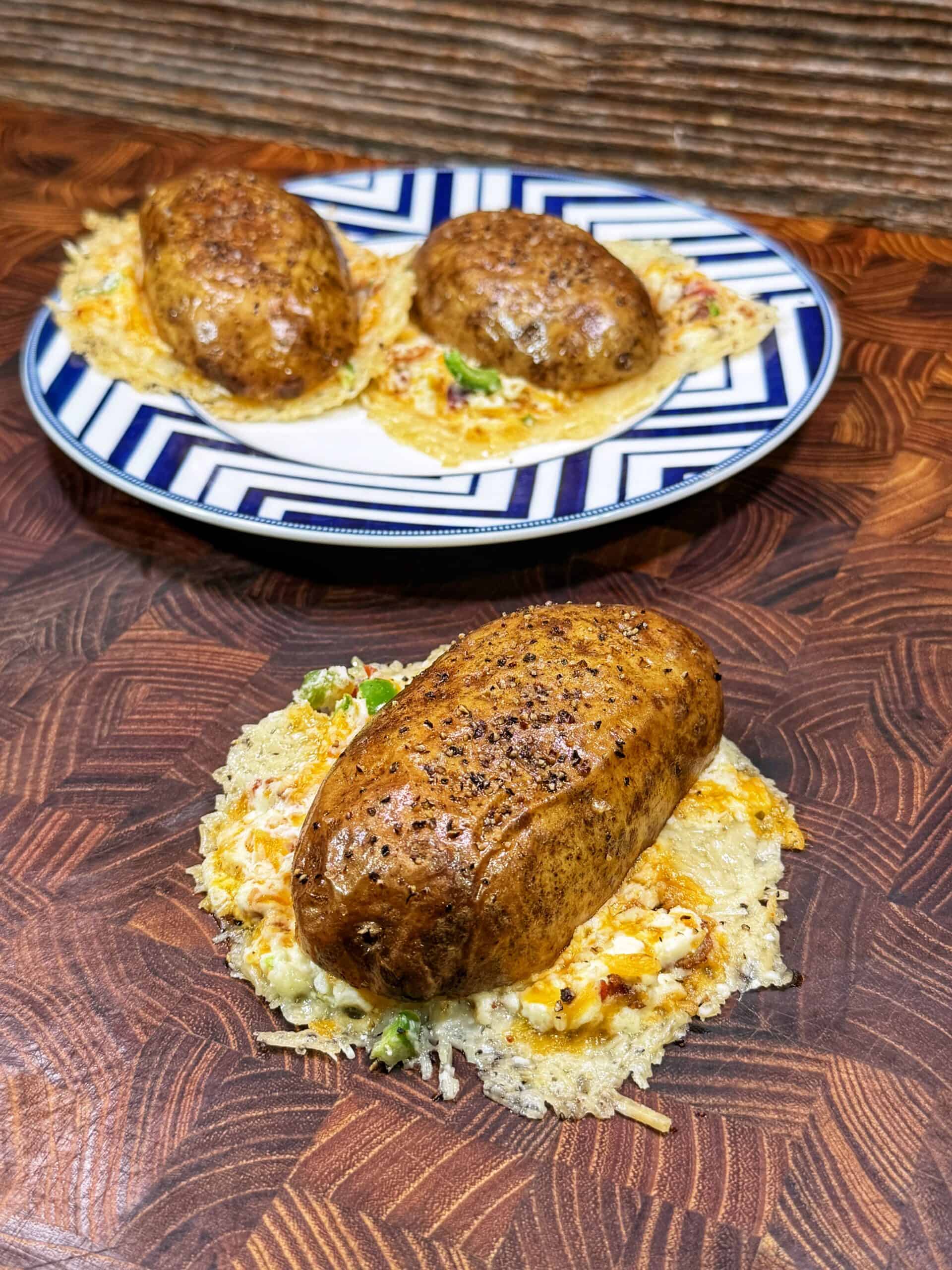Three baked potatoes topped with melted cheese and herbs, served on a patterned plate and a wooden surface. The potatoes rest on a layer of crispy, golden cheese.