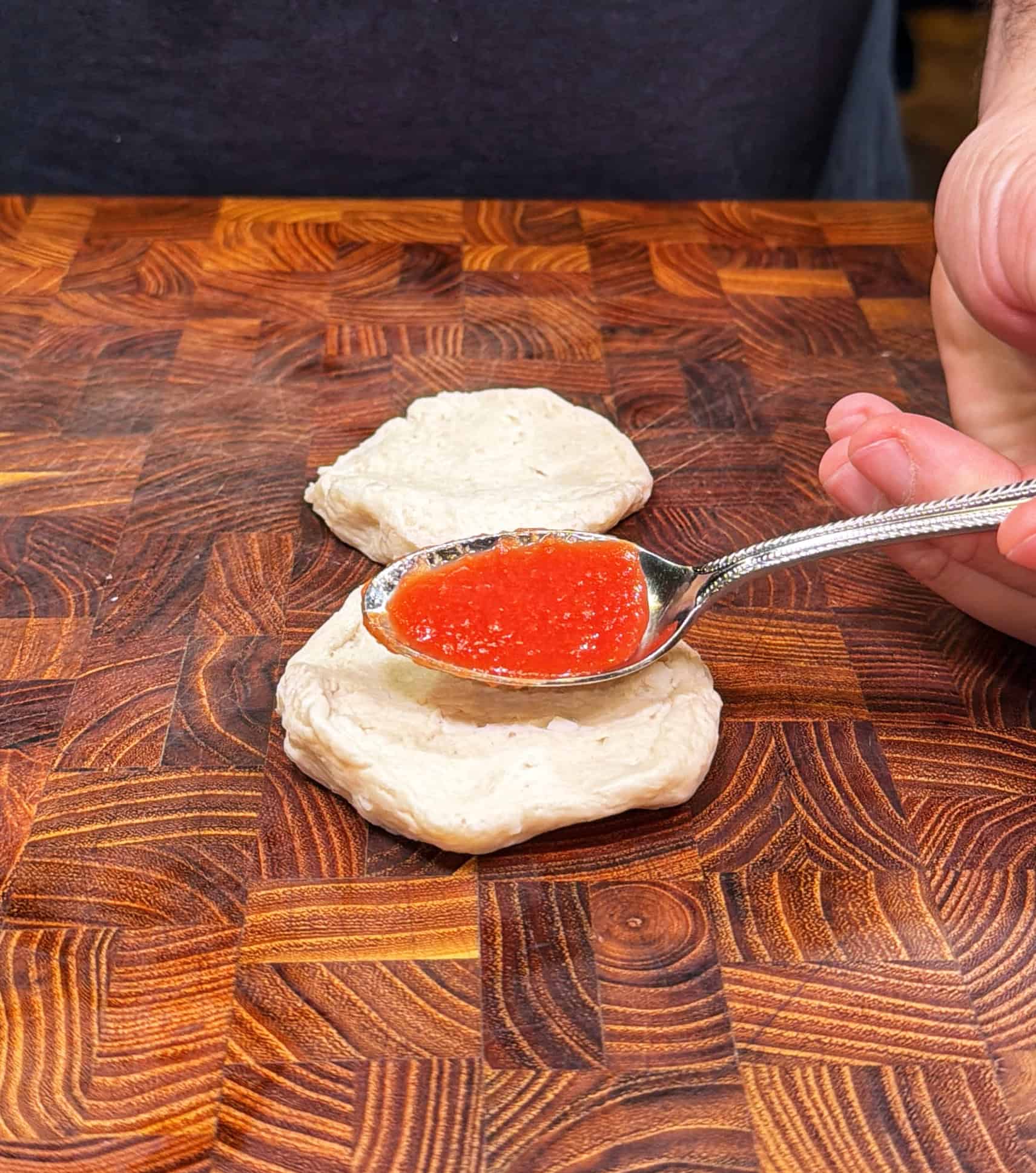 A hand holds a spoon with tomato sauce, spreading it onto a round piece of dough on a wooden cutting board, with another dough piece nearby.