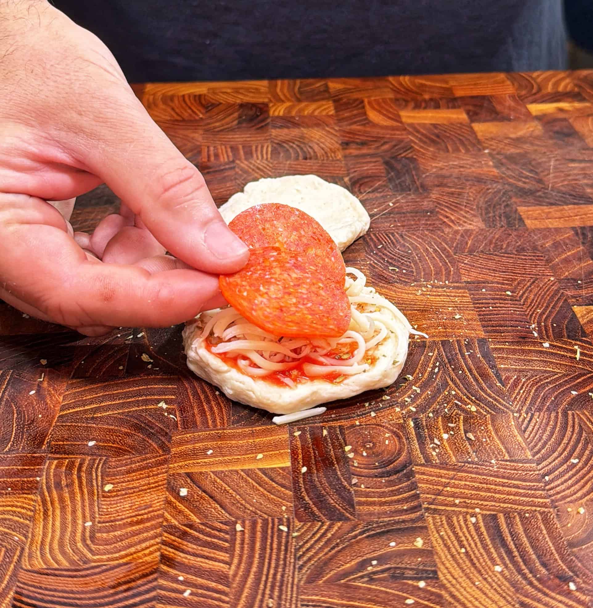 A hand places a slice of pepperoni on top of a small round dough with tomato sauce and shredded cheese, on a wooden cutting board.