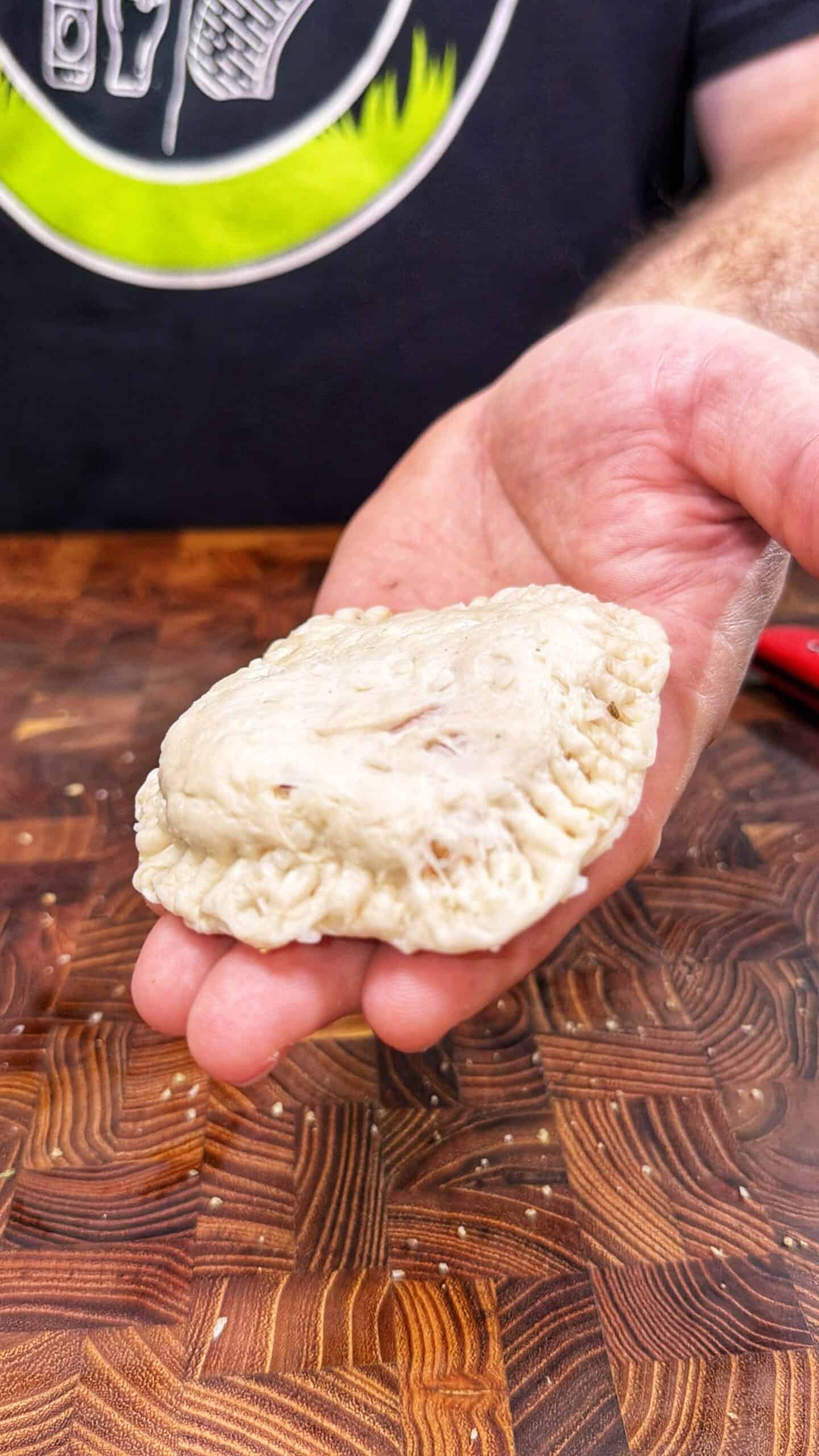 A person holds an uncooked, hand-crimped empanada or pastry over a wooden surface, wearing a dark shirt with a circular design.