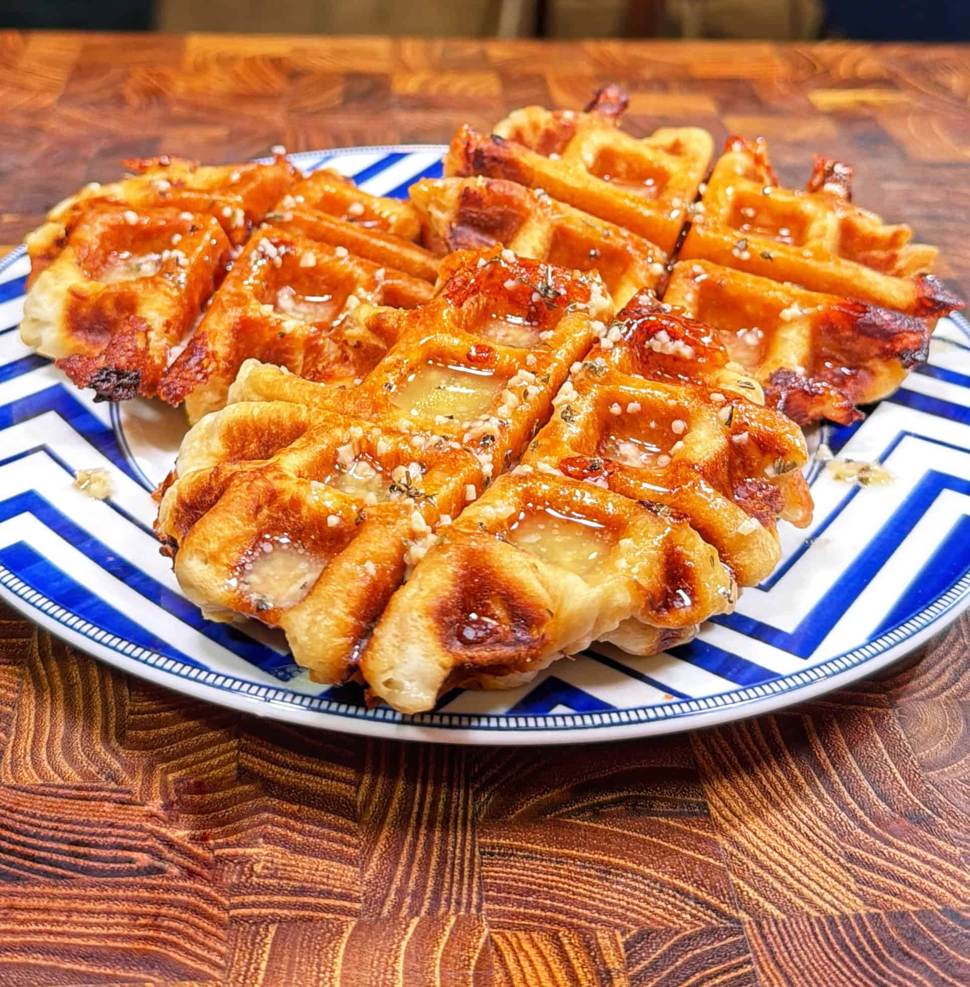 A plate with golden, crispy waffles drizzled with syrup and sprinkled with crushed nuts, placed on a wooden table. The plate has a blue and white zigzag pattern.