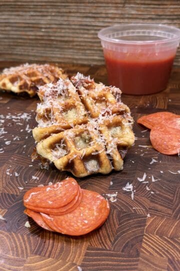 A waffle topped with grated cheese sits on a wooden board beside slices of pepperoni, more grated cheese, and a plastic container of red sauce, with another waffle in the background.