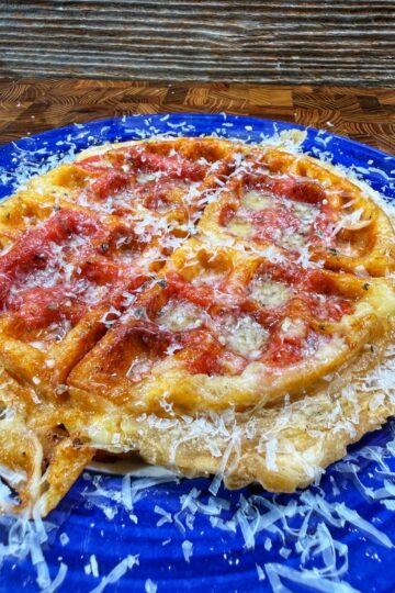 A golden waffle topped with tomato sauce and grated cheese sits on a bright blue plate, with extra cheese scattered around. The background is a rustic wooden surface.