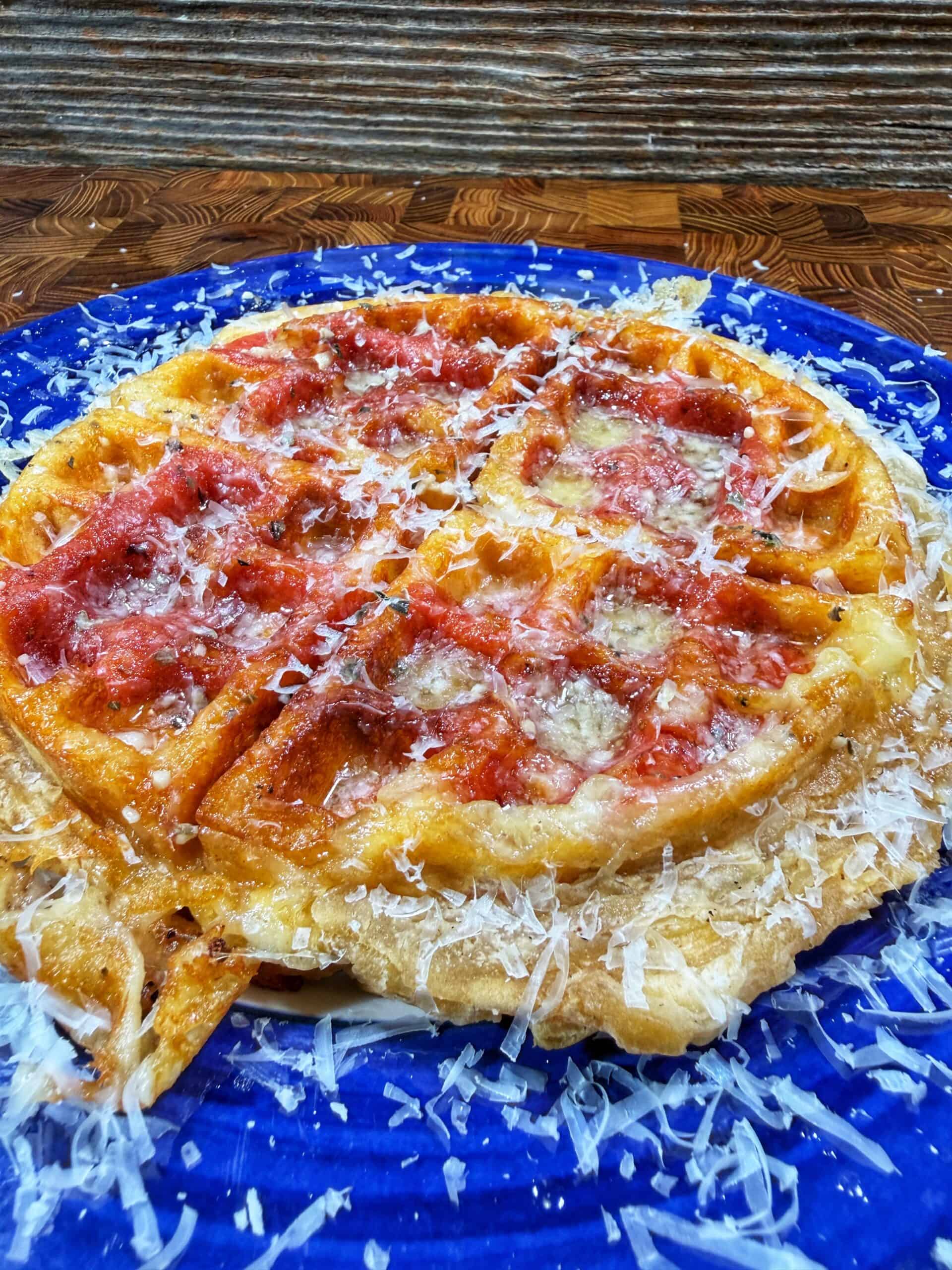 A golden waffle topped with tomato sauce and grated cheese sits on a bright blue plate, with extra cheese scattered around. The background is a rustic wooden surface.