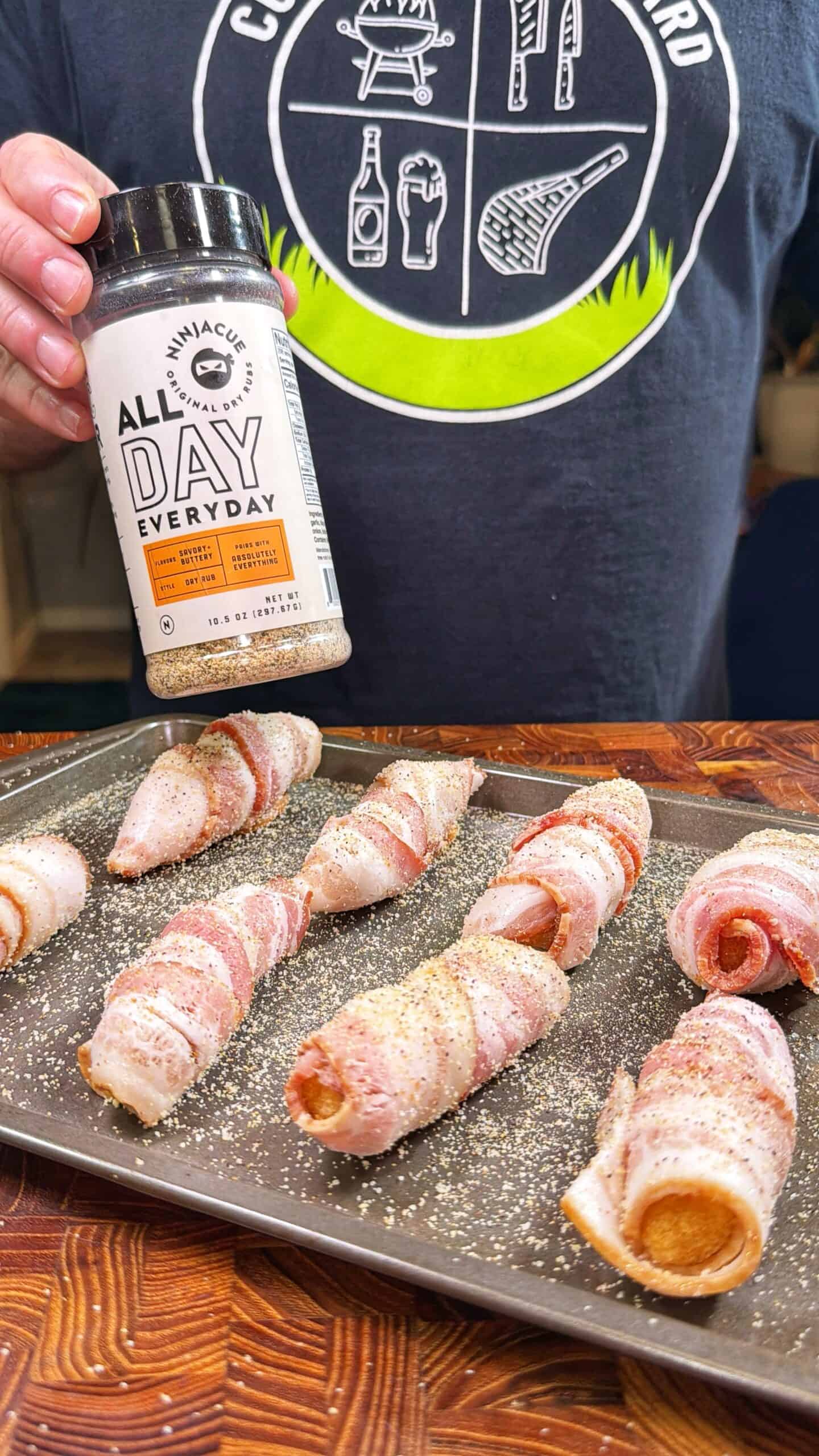 A person holds a jar of seasoning labeled “All Day Everyday” over a tray of hot dogs wrapped in bacon, ready for cooking. The person wears a dark shirt with a graphic outdoor-themed design.
